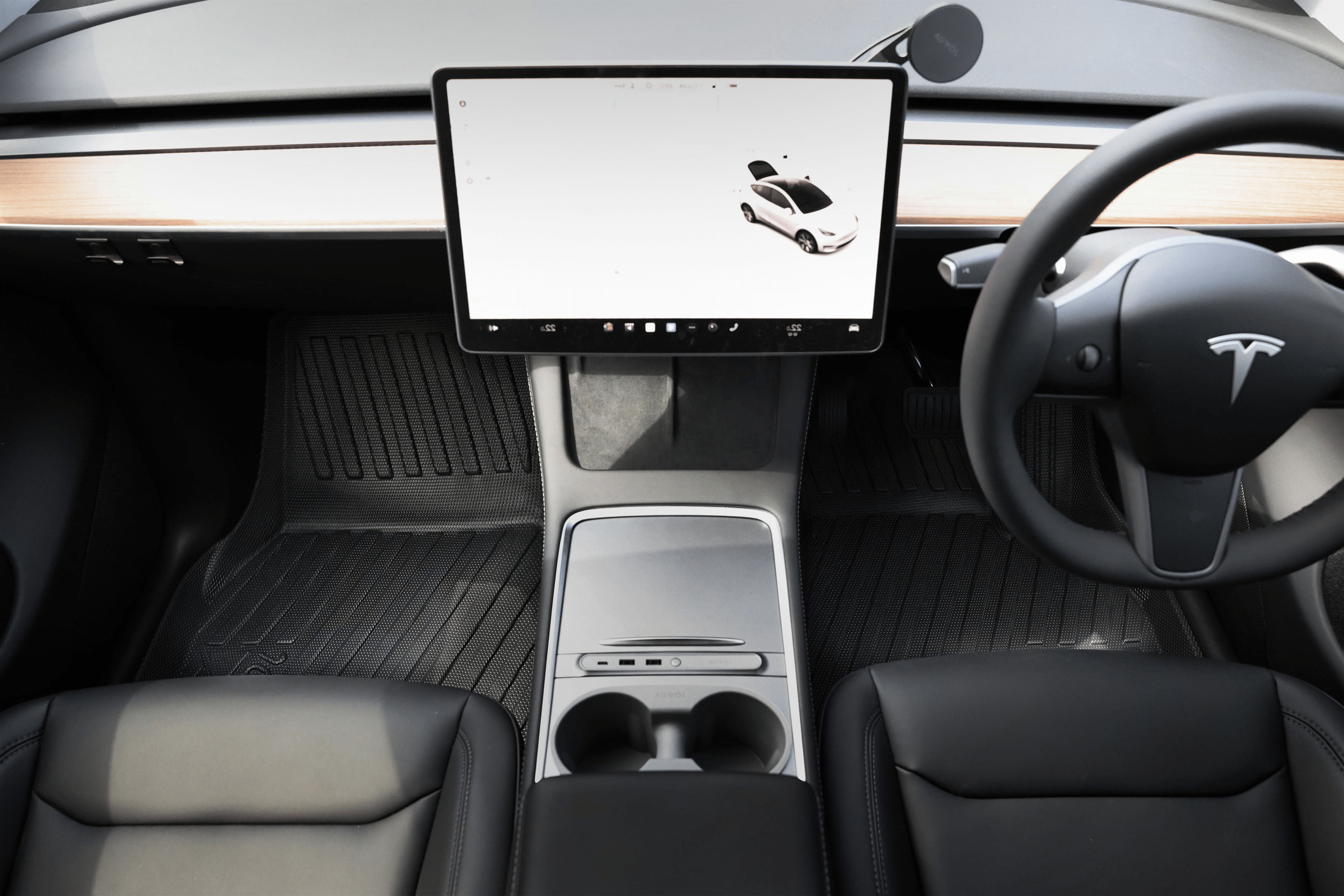 Interior of a Tesla car showing the steering wheel, large central touchscreen display, minimalist dashboard, cup holders, and black leather seats. The display shows an image of the car.