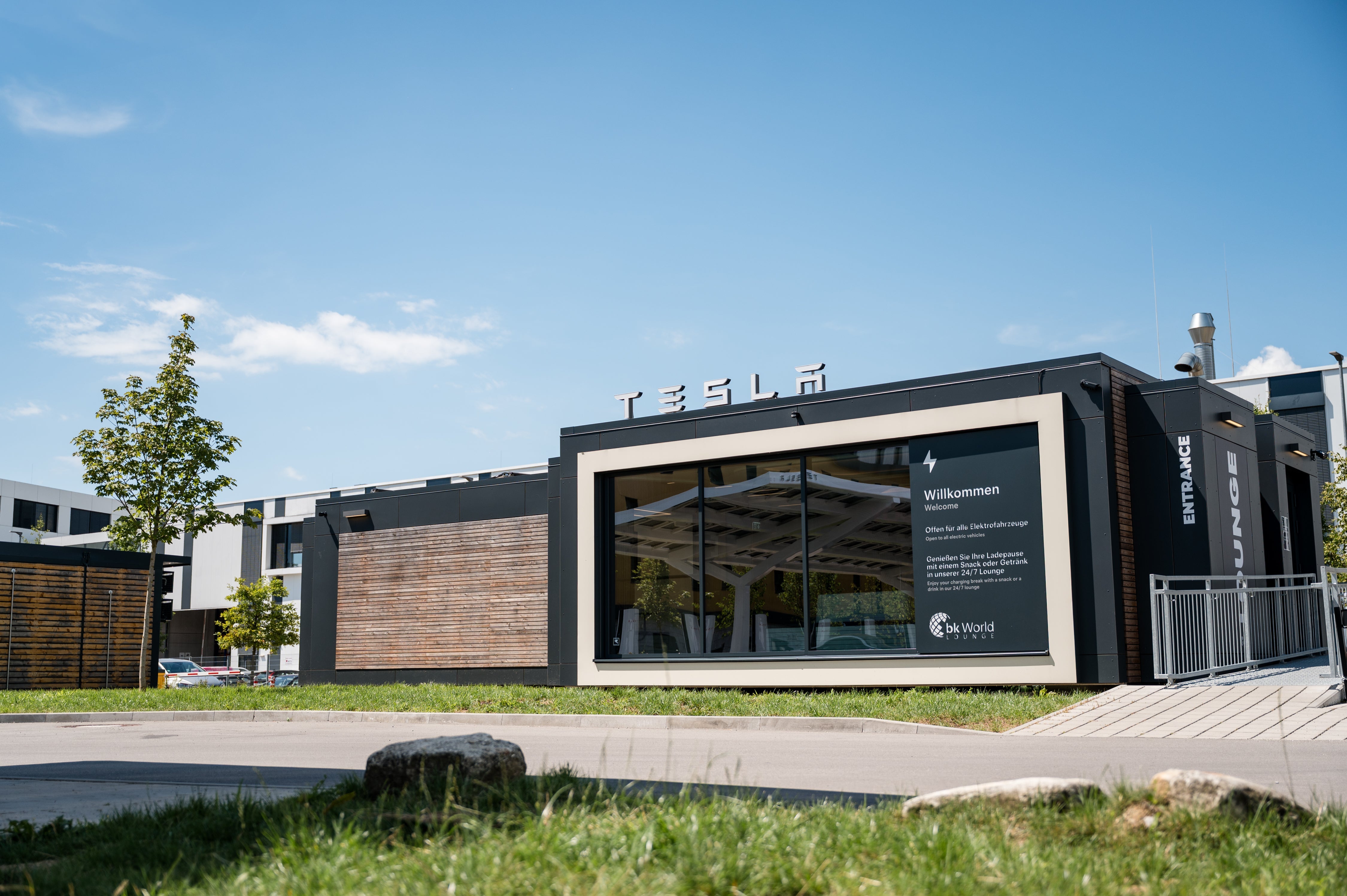 Modern building with large glass window and TESLA sign on top, surrounded by greenery under a clear blue sky. A sign on the window says "Willkommen" and "Entrance.