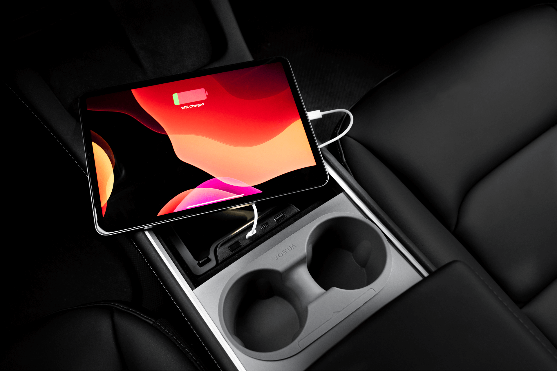 A tablet with a low battery warning is charging on a car's center console between two cup holders, with a cable attached.