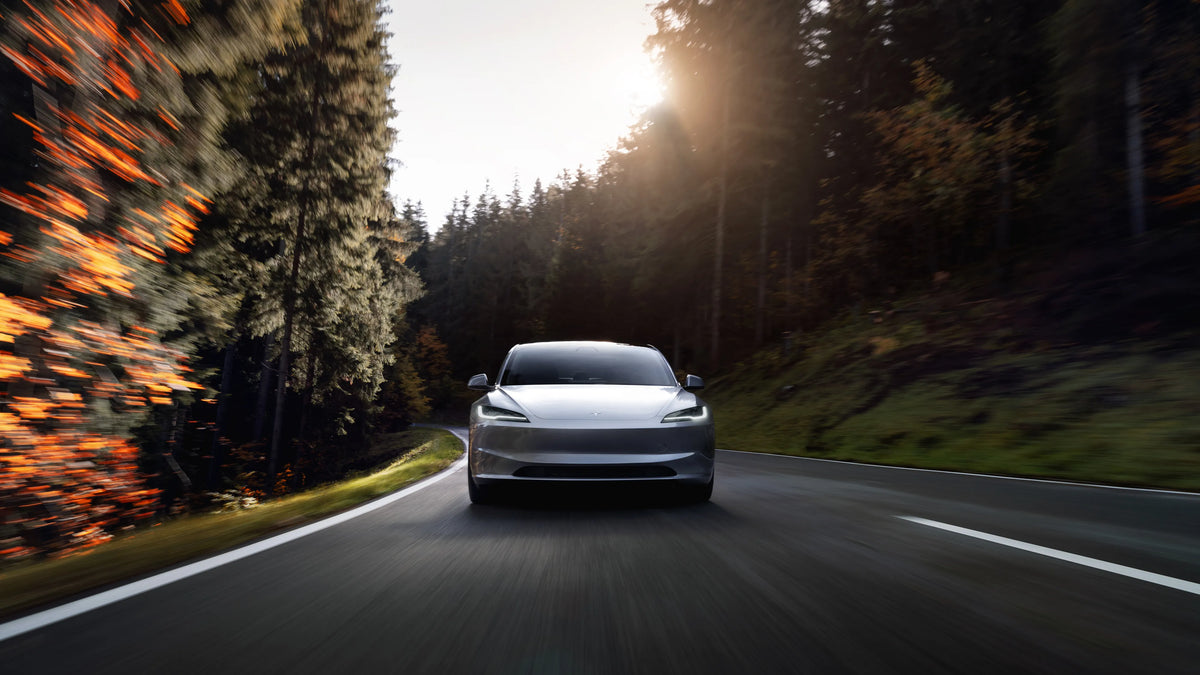 A silver Tesla Model 3 drives along a winding road through a forest at sunset.