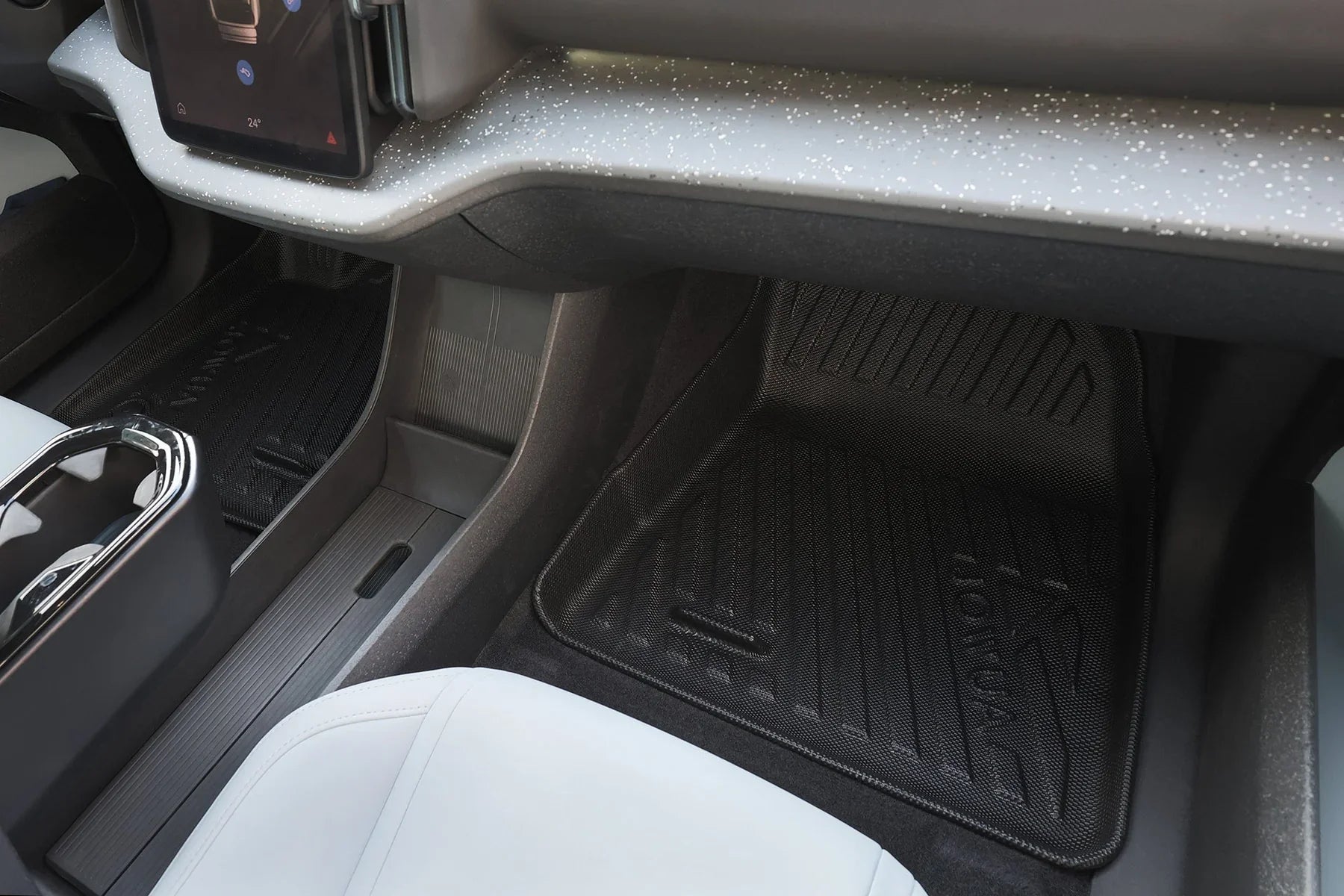 Close-up of a car’s front passenger footwell, showing a textured dashboard, black floor mat with "HONDA" branding, light-colored seat, and part of the center console with a touchscreen above.