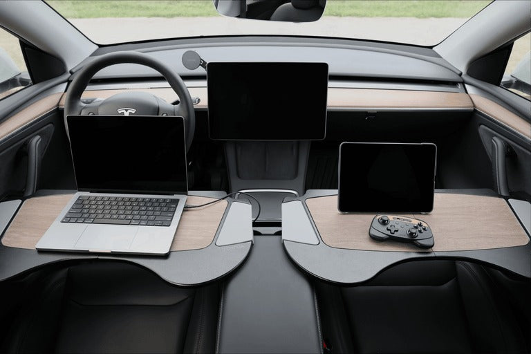 A Tesla car interior with two desks; the left holds a laptop, the right holds a tablet and game controller. Two large screens are mounted on the dashboard, and the steering wheel is visible.