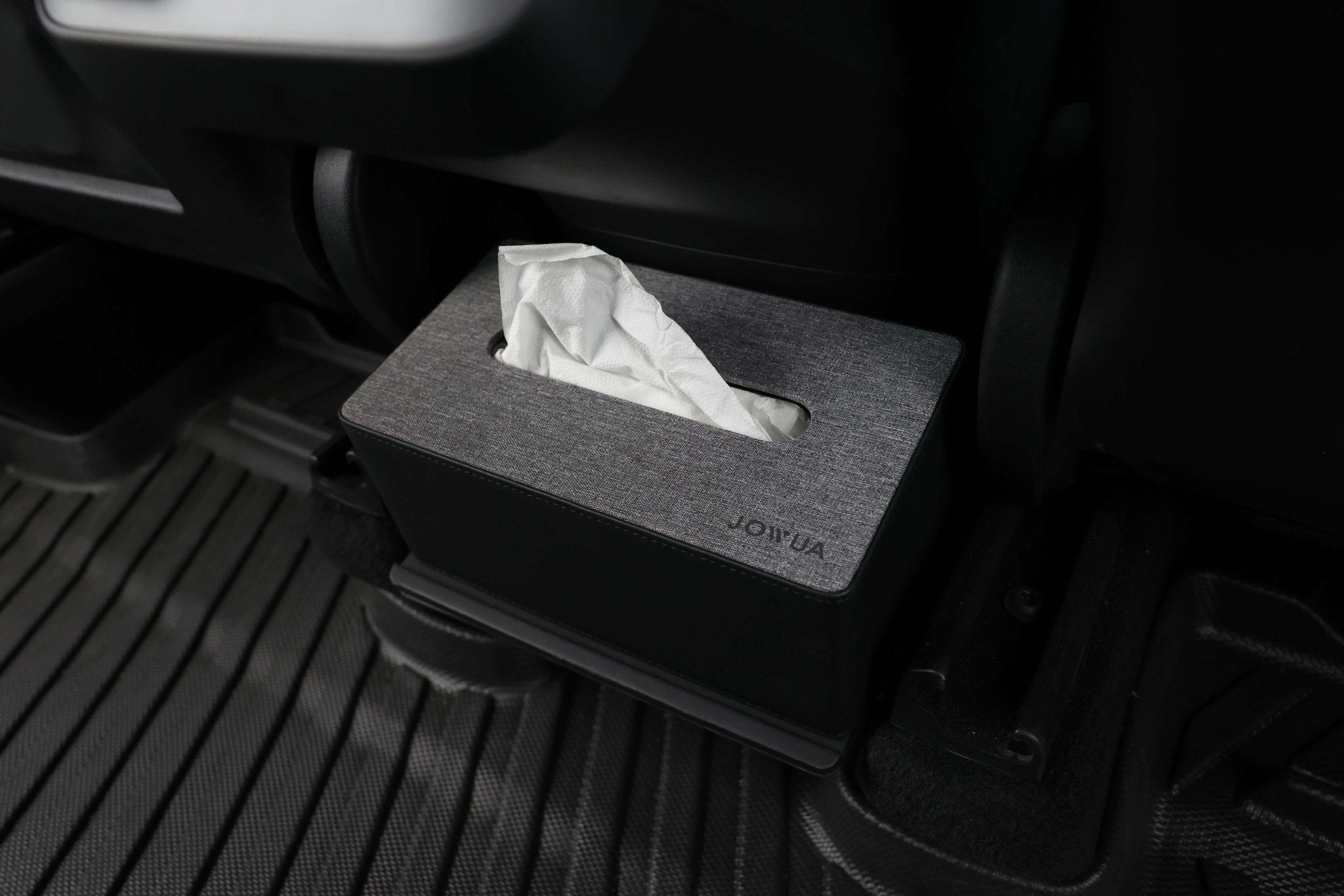 In the Tesla Model Y's footwell area, a sleek JOWUA Model Y Tissue Box Holder Set with gray fabric covering rests on a textured, dark floor mat. The box is partially opened, with a white tissue peeking out, adding both style and convenience to your car's interior.