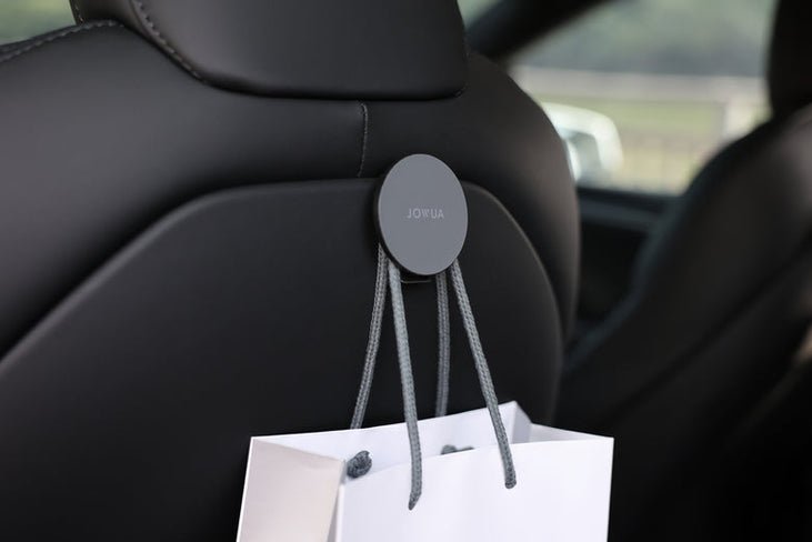 A white shopping bag hangs from gray cords hooked to a Tesla Model Y Juniper (2025+) seat, showcasing JOWUA’s Premium Interior Essentials Set against a blurred car window and greenery backdrop.