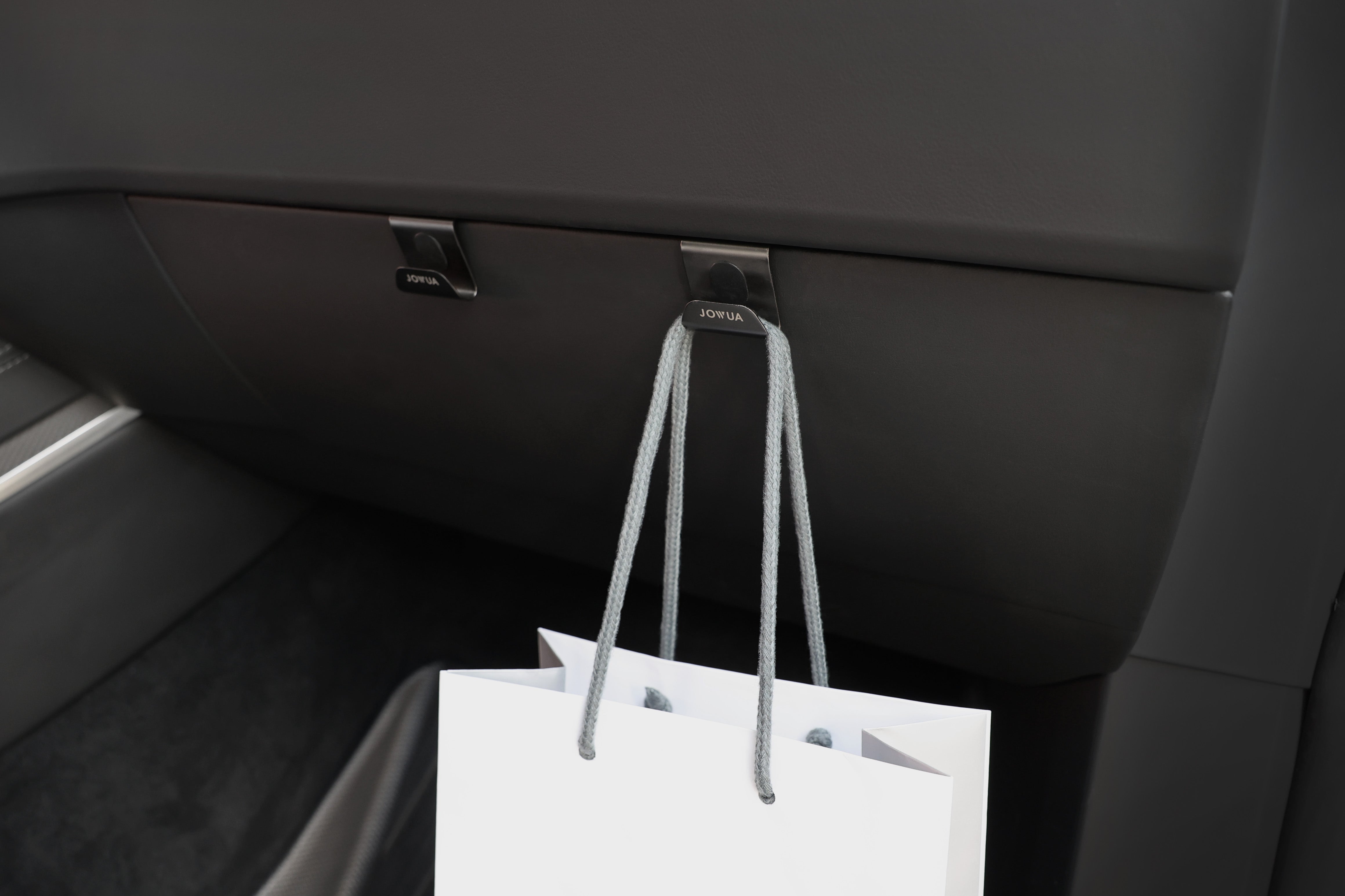 A JOWUA Glovebox Hook for Tesla Model S/X/3 Highland 2024+/Y Juniper 2025+ holds a white shopping bag with gray handles inside the car, enhancing the dark interior.
