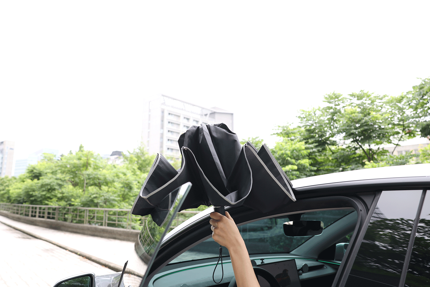 A person holds a JOWUA Type-C Electric Umbrella, black and reverse folding, while standing next to an open car door on a cloudy day with trees and buildings in the background.