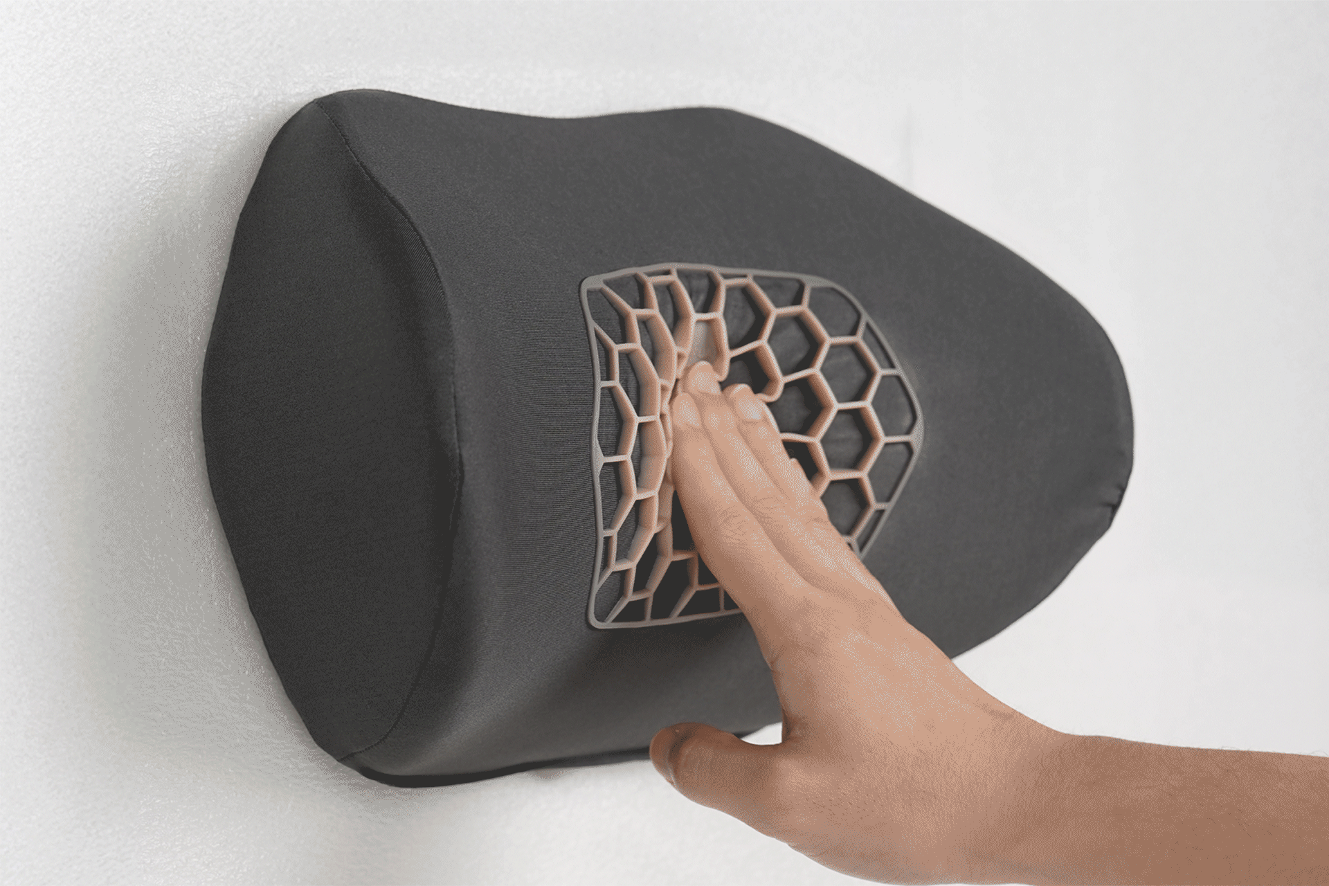 A hand presses into the JOWUA Tesla Neck Pillow, an ergonomic gray cushion with a hexagonal mesh center, mounted on the wall.