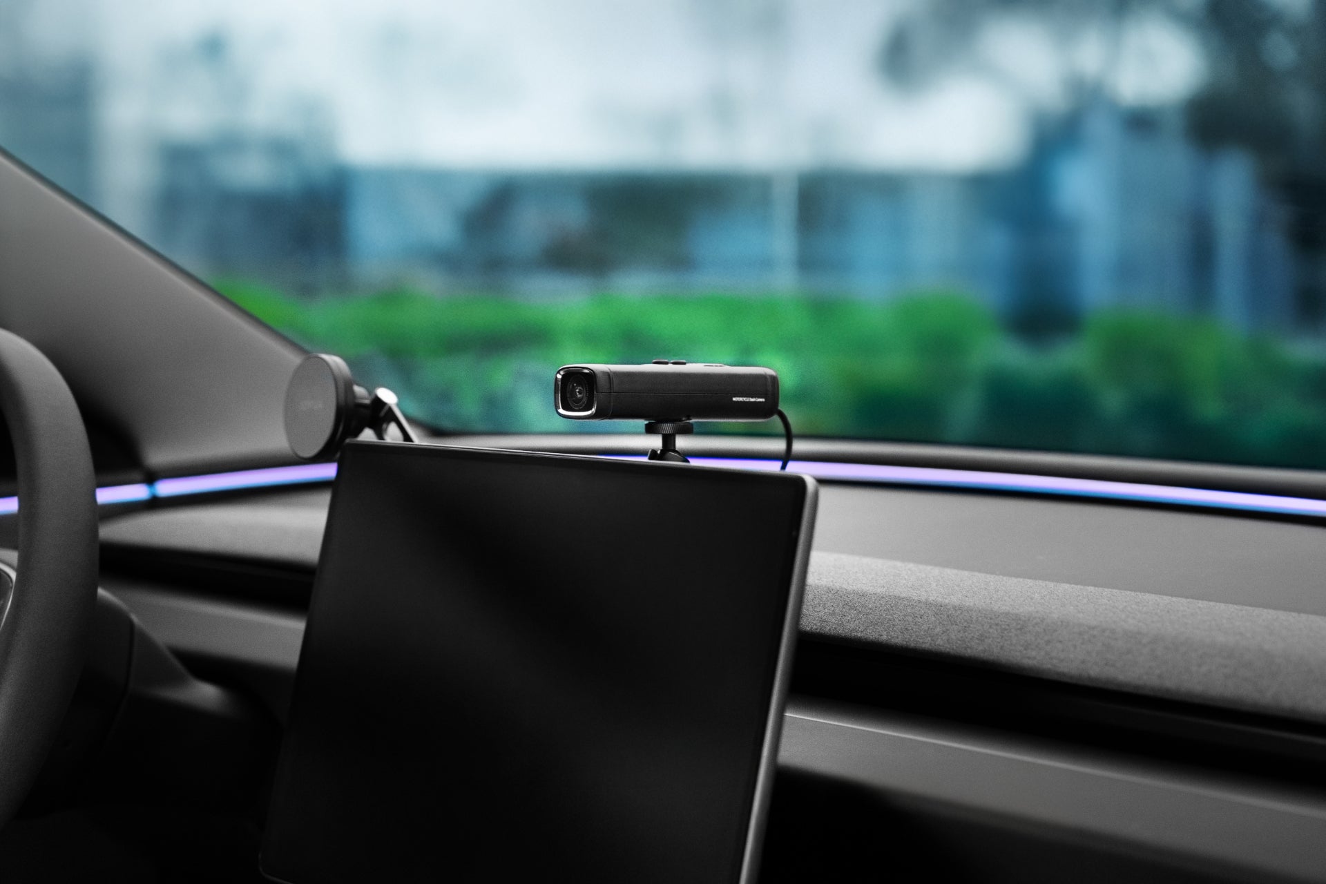 A JOWUA 360° Multi Function Mount holds a small black webcam on top of a tablet inside a car, with greenery and buildings blurred through the windshield.