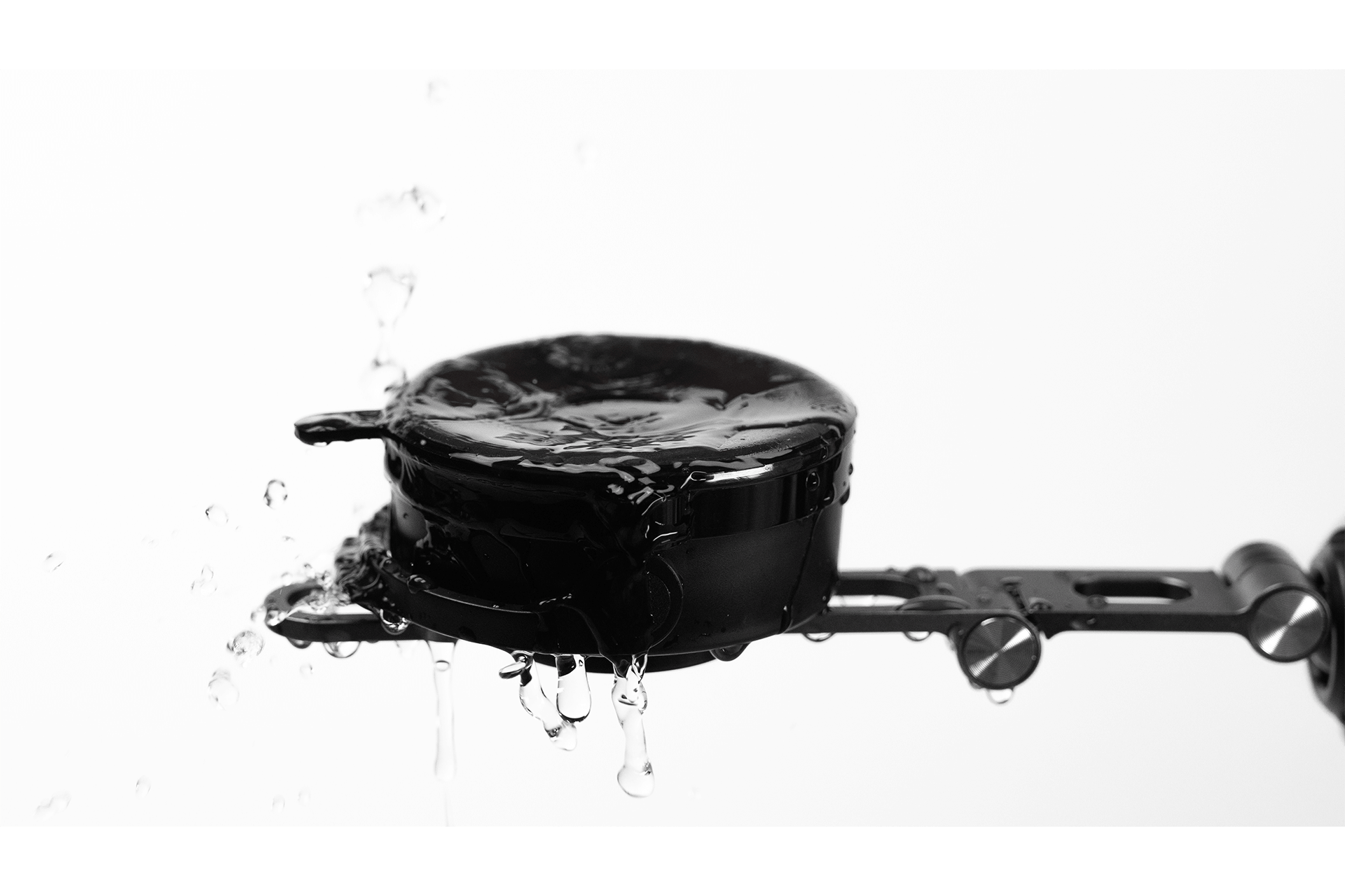 A black, round JOWUA Active Cooling Tesla Compatible Phone Mount (S/3/X/Y/Cybertruck) is splashed with water, droplets cascading down its sleek sides as it sits mounted on a metal arm against a plain white background.