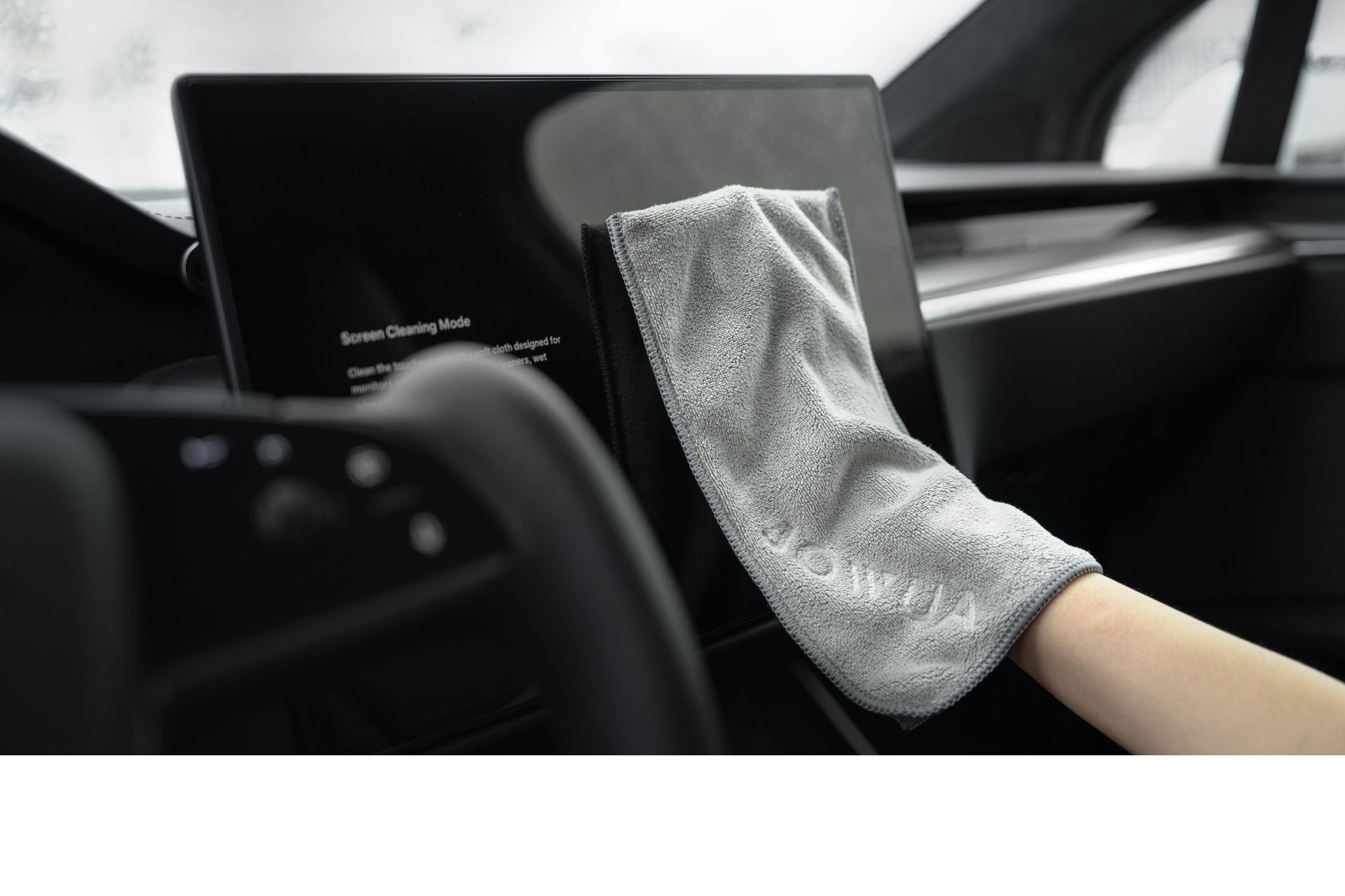 A hand is using a JOWUA Microfiber Cleaning Cloth, part of the Under Seat Collapsible Organizer & Microfiber Cleaning Cloth Combo, to clean a large touchscreen display inside a car. The interior is modern and sleek, with the steering wheel partially visible in the foreground.