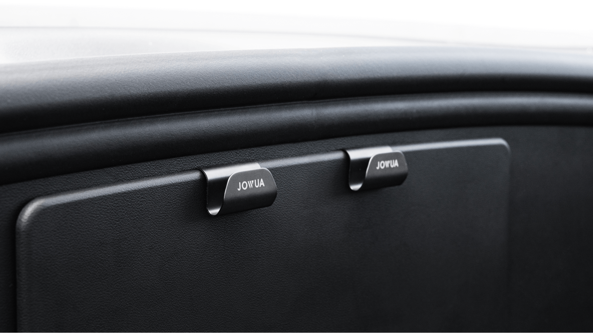 Close-up of a black glove compartment with two latches labeled "JOWUA" in white, set against a sleek dashboard—ideal for the Premium Interior Essentials Set (RHD) – Model Y Juniper (2025+) from JOWUA.