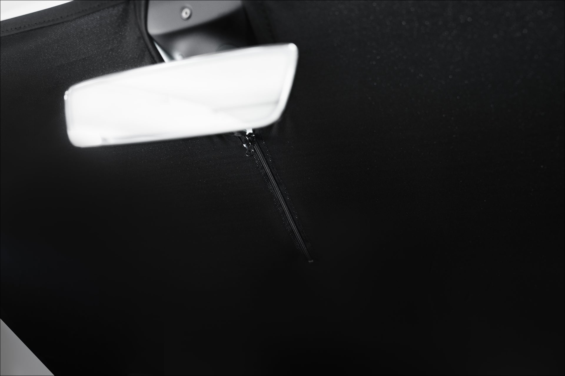 Close-up of a JOWUA Model Y Ultimate Sunshade Bundle interior, emphasizing the rearview mirror and a partially unzipped black fabric roof cover. The focus is on the zipper and the contrasting textures of the mirror and Dark Matter mesh fabric, which provides UV protection.