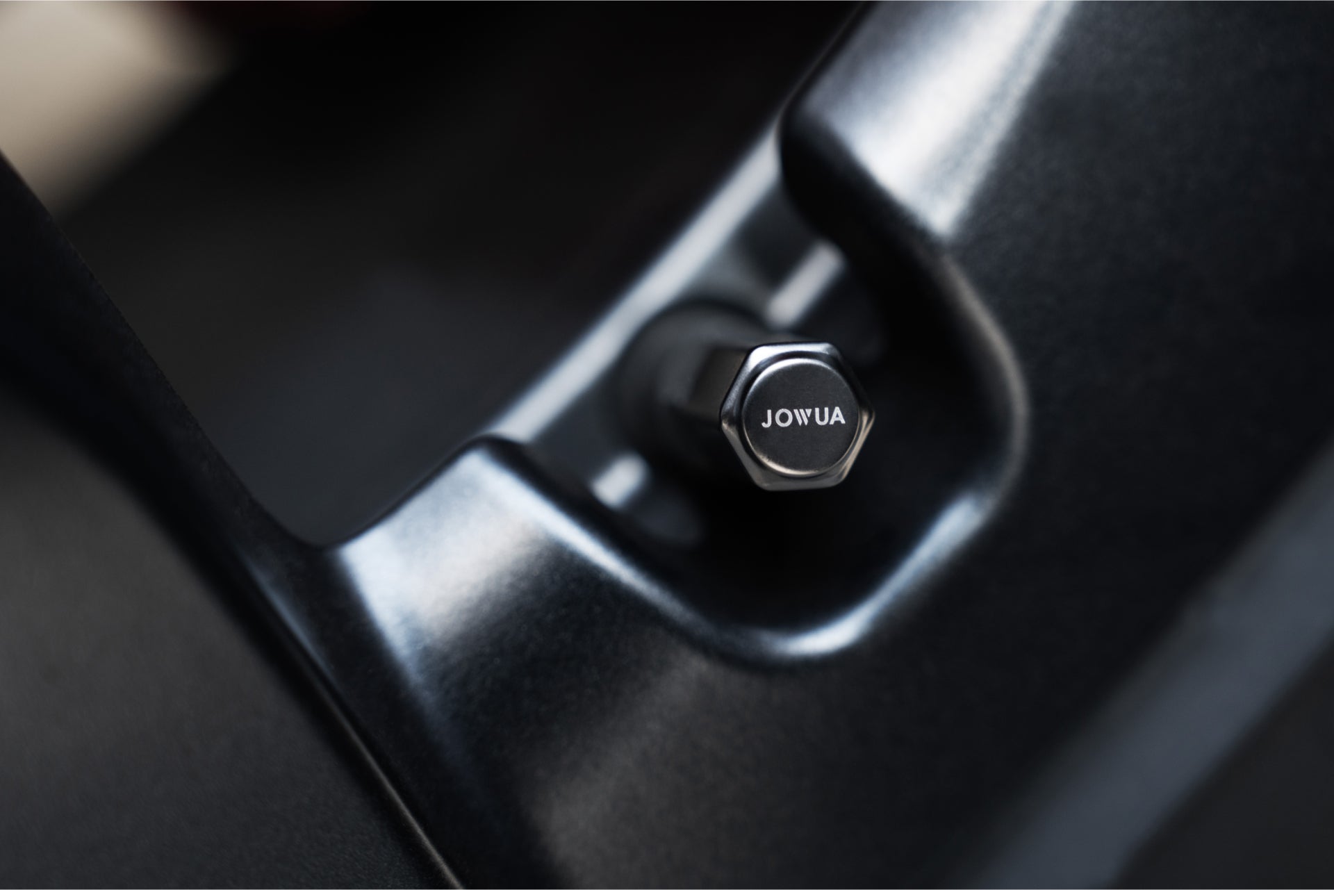 A close-up of a metallic hexagonal tire valve cap from JOWUA, crafted from durable zinc alloy and snugly placed within a sleek black holder. The polished surface of the cap contrasts with the matte finish, ensuring airtight leak protection for optimal performance.