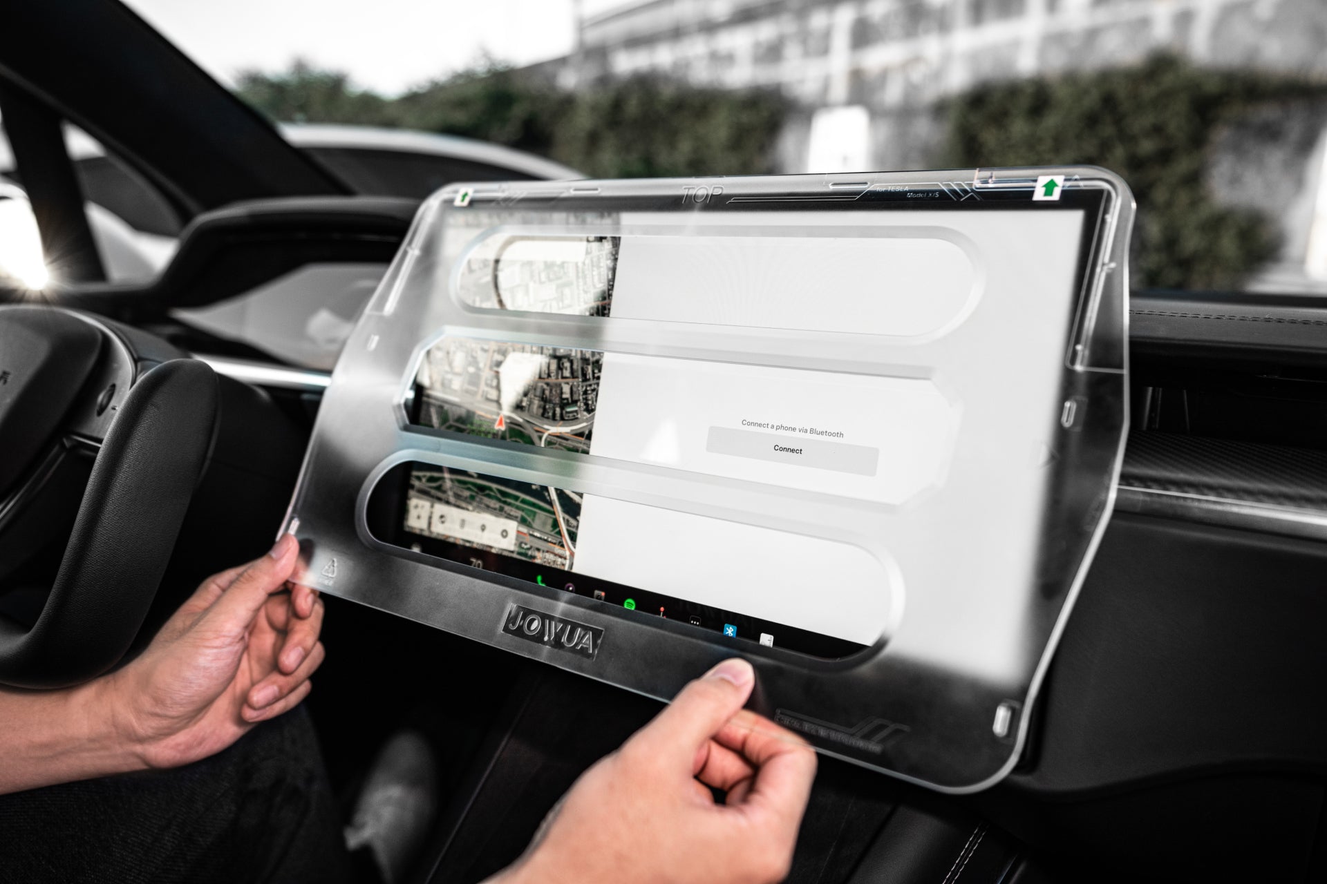 Hands holding a cutting-edge Tempered Glass Screen Protector (Model S/X) by JOWUA inside a car, displaying a map application and connection prompt on its matte surface. The interior showcases sleek, modern design elements with a dashboard in the background, ensuring an anti-fingerprint display for smooth navigation.