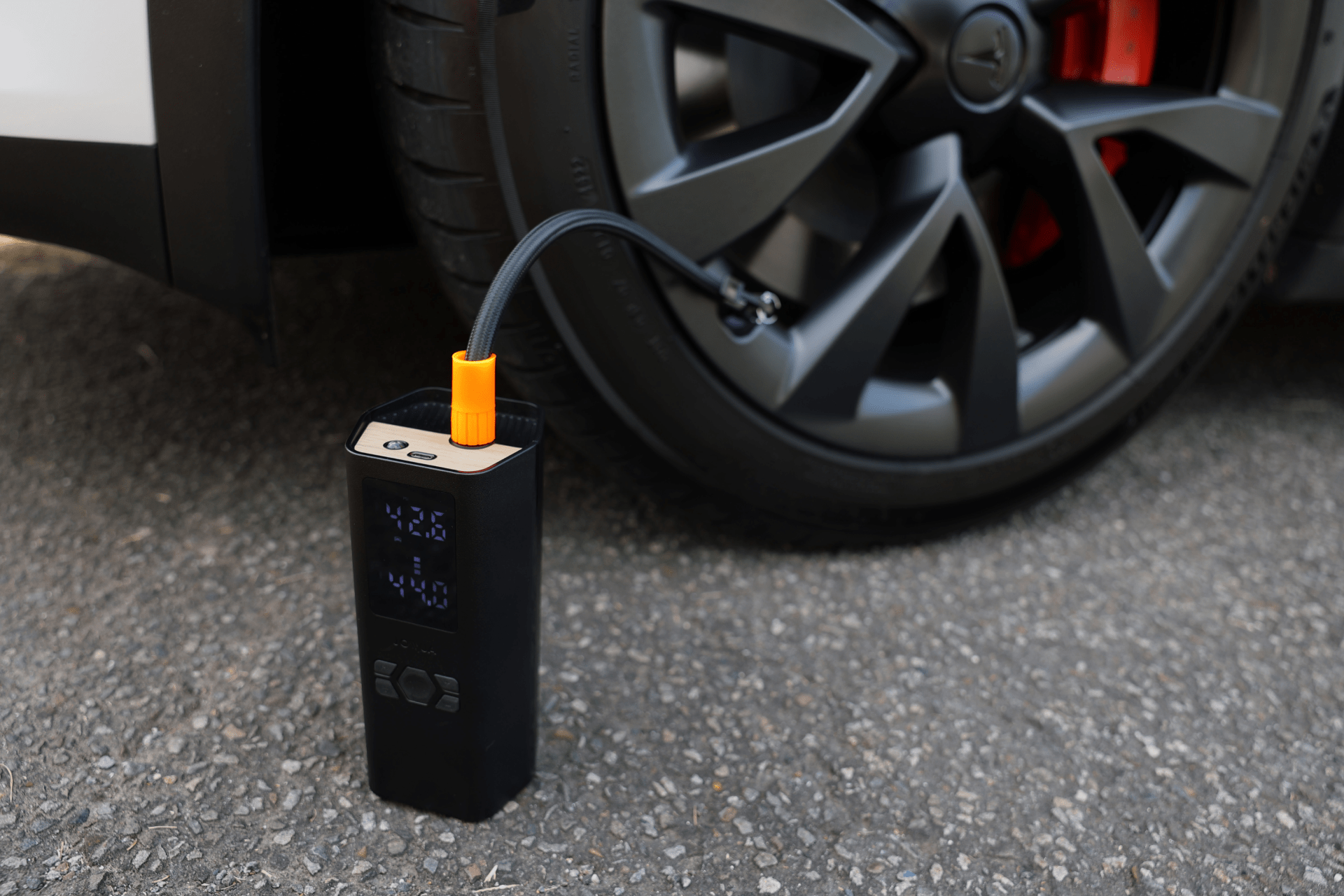 The JOWUA Portable Air Compressor + Charging Solution Kit is inflating Tesla tires on a paved surface, displaying digital readings on its screen and connecting securely to the tire valve with its attached hose.