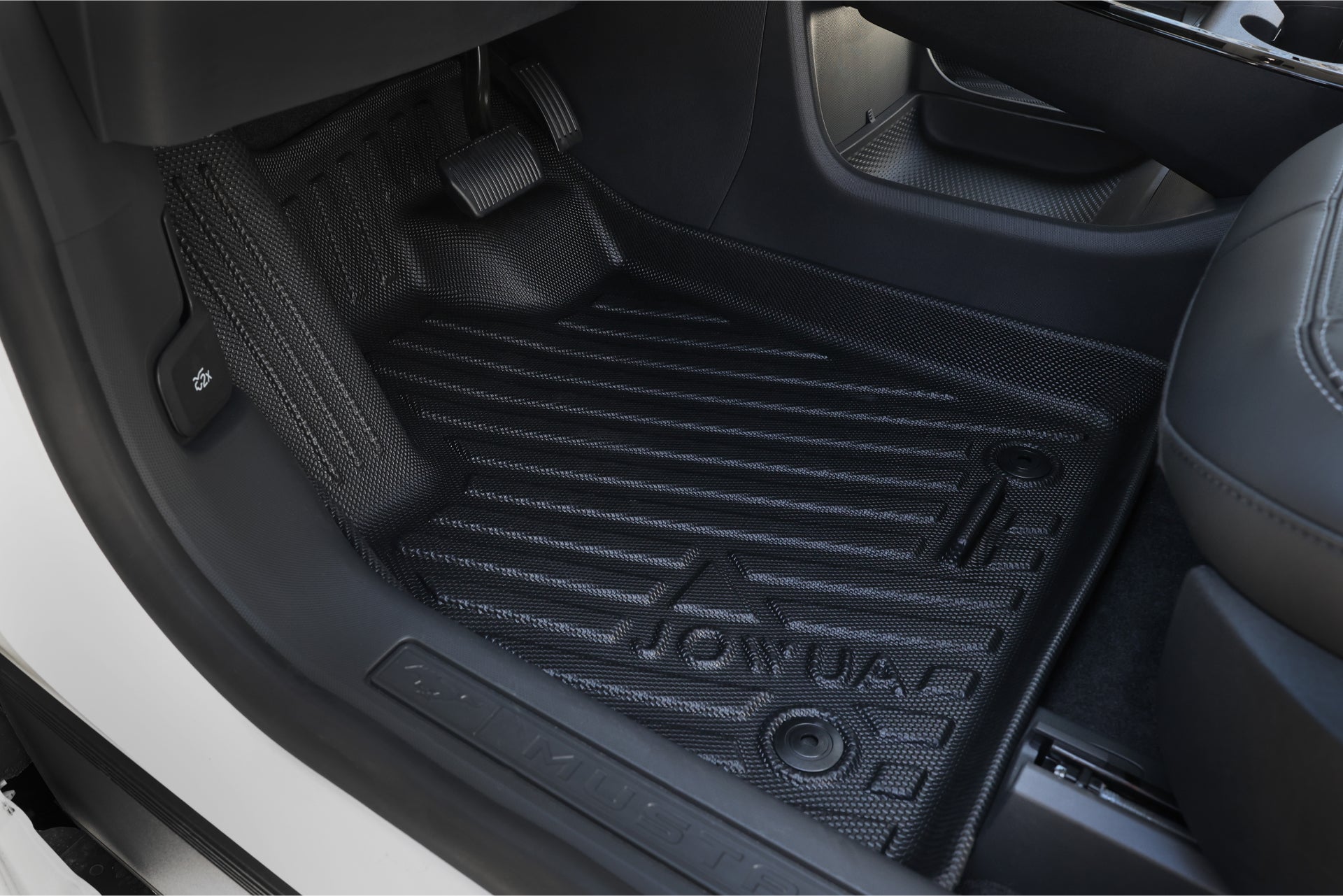 A close-up view of a car's interior showcases a black, textured all-weather liner by JOWUA for the Ford Mustang Mach-E. The liner protects the area beneath the accelerator and brake pedals, which are visible in the image, with the seating area partially visible on the right.