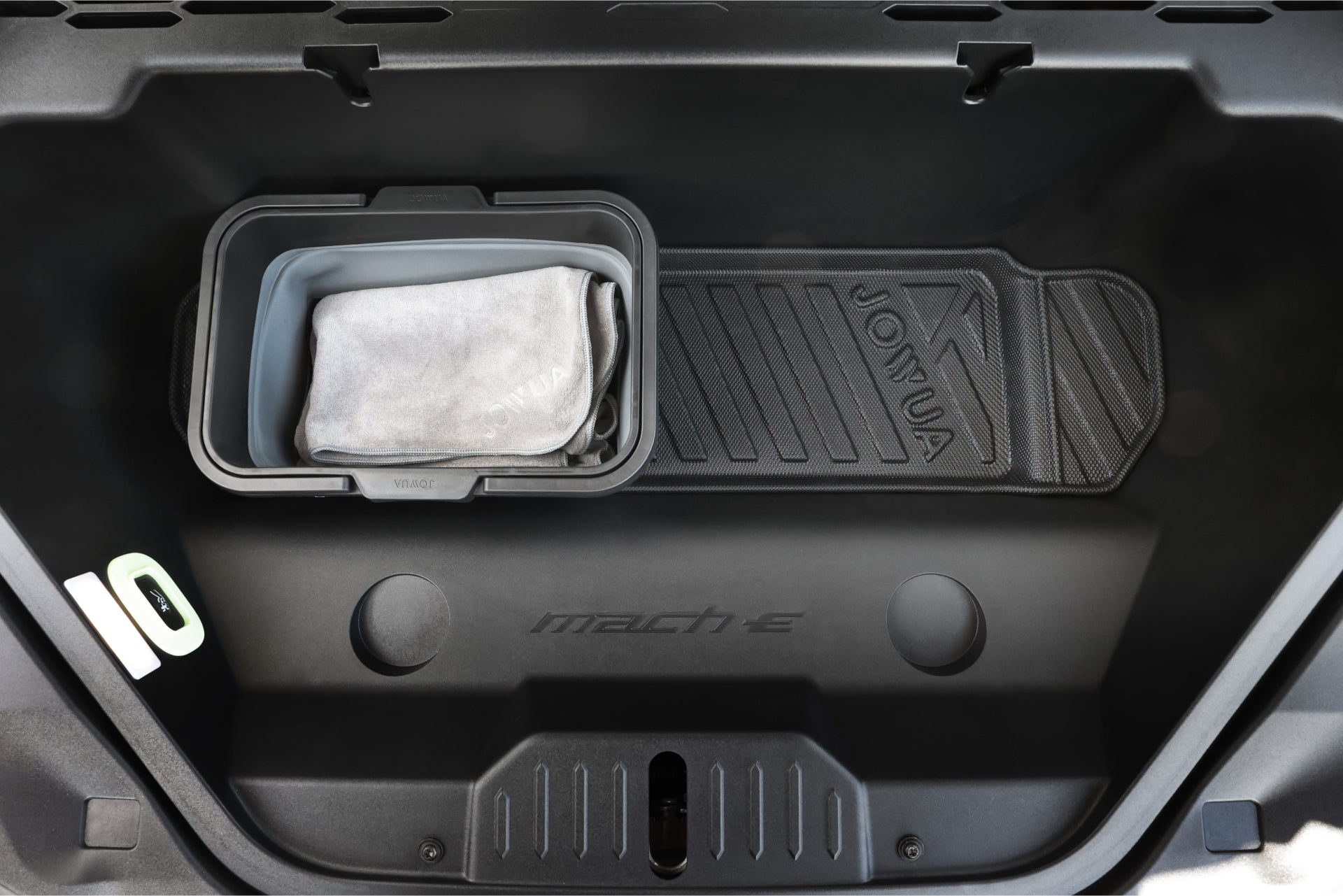The open car trunk reveals a storage bin containing a folded gray cloth. A black, textured floor mat with the "JOWUA" brand is placed on the All Weather Trunk Liner for the Ford Mustang Mach-E, with "Mach-E" engraved in the trunk to emphasize its thoughtful design.