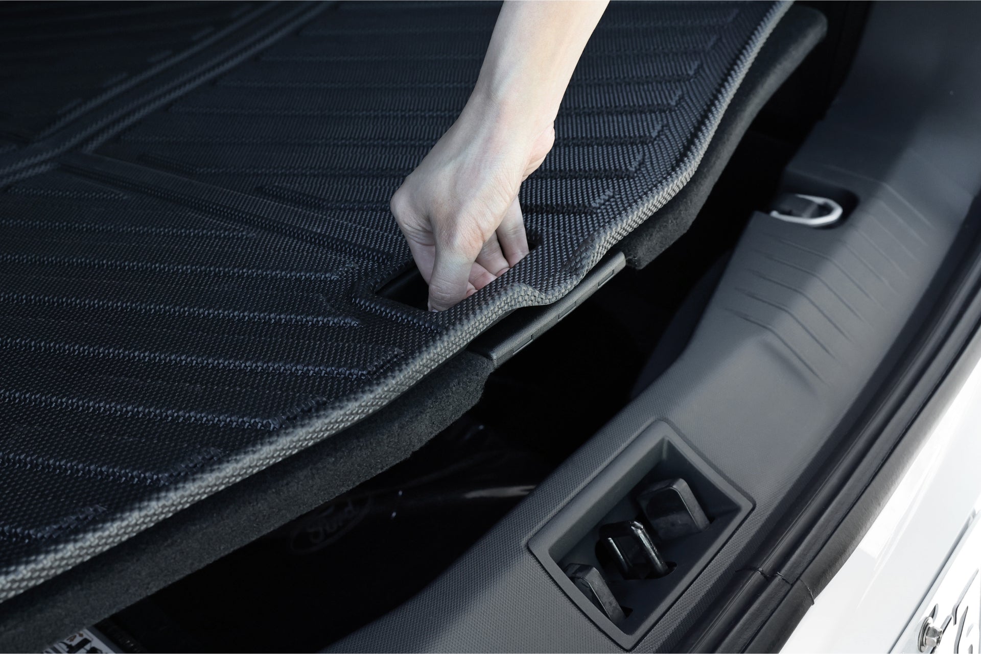 A hand raises the trunk cover of a Ford Mustang Mach-E, showcasing the sleek storage compartment beneath. The car's interior looks clean and modern, featuring a textured surface on the cover, enhanced by JOWUA's All Weather Trunk Liners—ideal for any adventure.