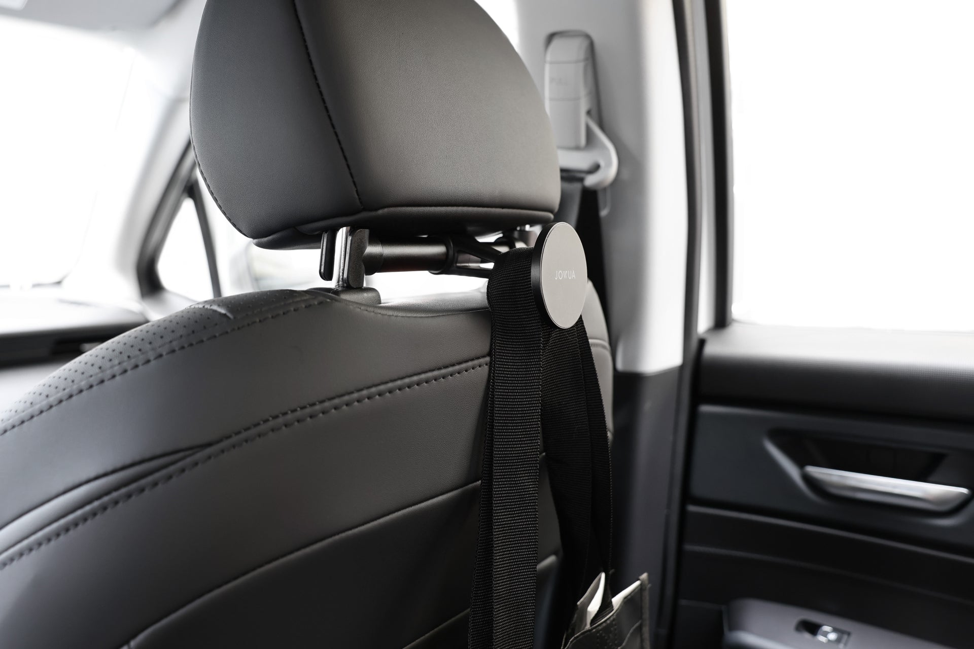 In the car's interior, the focus is on the back of the front seat headrest, where a JOWUA Universal MagSafe Car Seat Holder with an anti-slip design is securely attached to hold a hanging item. The seat features black leather upholstery, and a magazine is partially visible in the pocket.