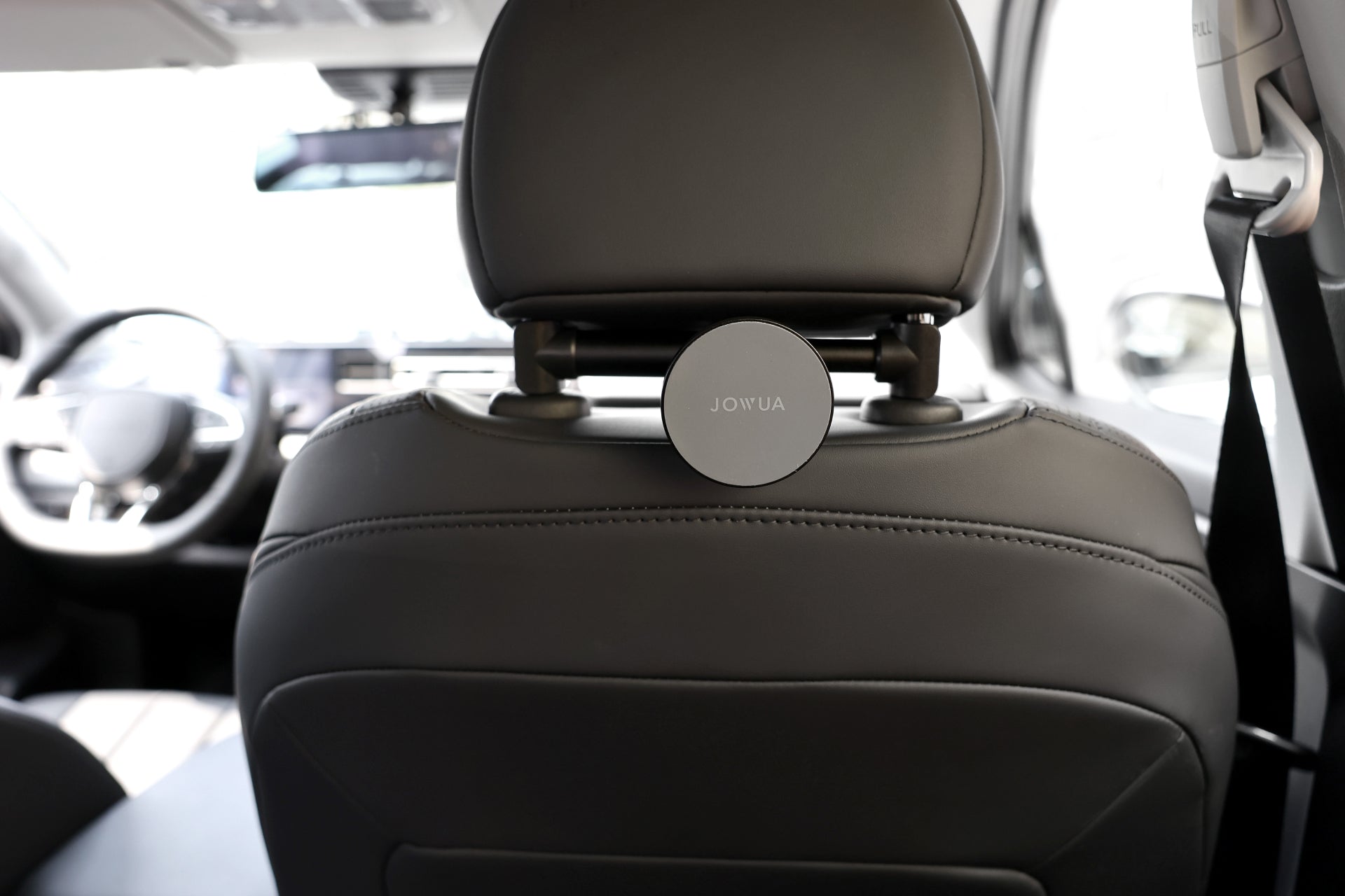 Close-up of a car's black leather front seat with a Universal MagSafe Car Seat Holder by JOWUA attached to the headrest. The car's interior, showcasing an anti-slip design dashboard and steering wheel, is slightly visible in the background.