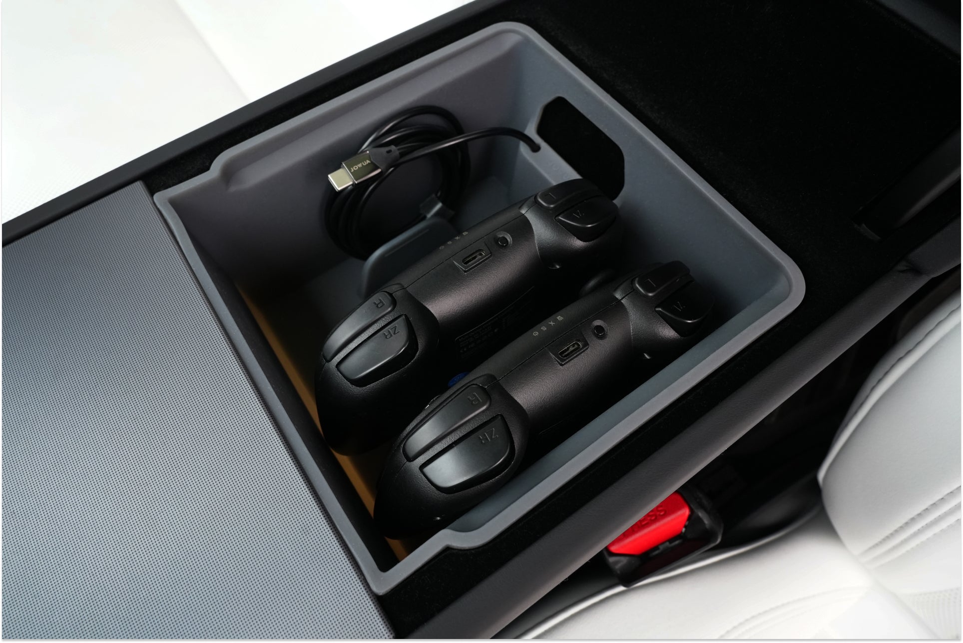 A close-up of the JOWUA Armrest Organizer Set for Tesla Model Y Juniper 2025+ / Model 3 Highland 2024+, holding two black wireless game controllers and a USB charging cable neatly stored for easy access.