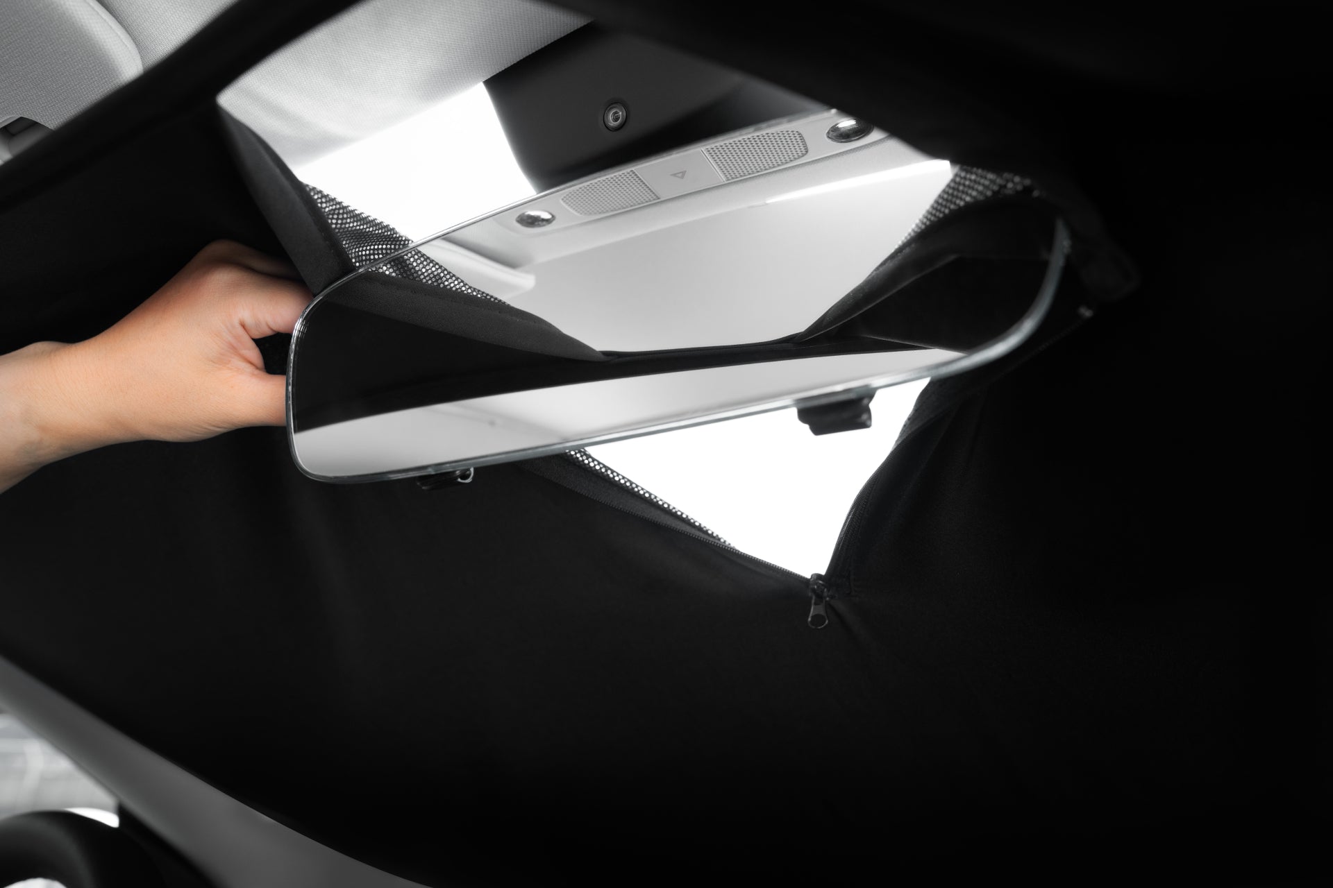 A person adjusts the rearview mirror of a Tesla Model Y, partially covered by the black, stitched JOWUA Model Y Ultimate Sunshade Bundle. The fabric provides UV protection while still allowing full adjustability of the mirror. In the background, the car's interior showcases its sleek design elements.