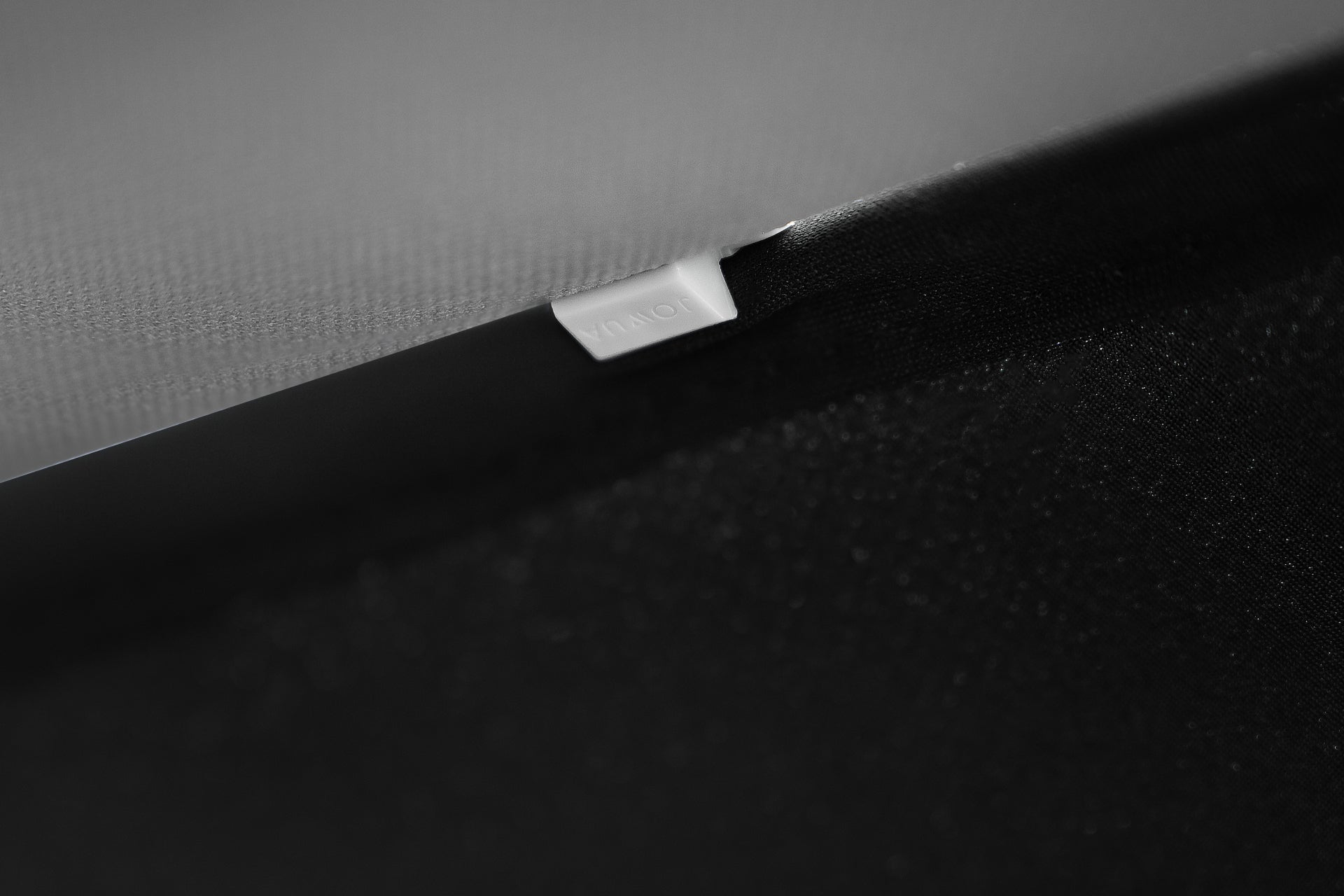 Close-up of a black and gray textured surface, featuring a small metallic tag engraved with "JOWUA." The image highlights the sleek design of the Model Y Ultimate Sunshade Bundle, reminiscent of Tesla Model Y craftsmanship, demonstrating attention to detail and innovative material use.