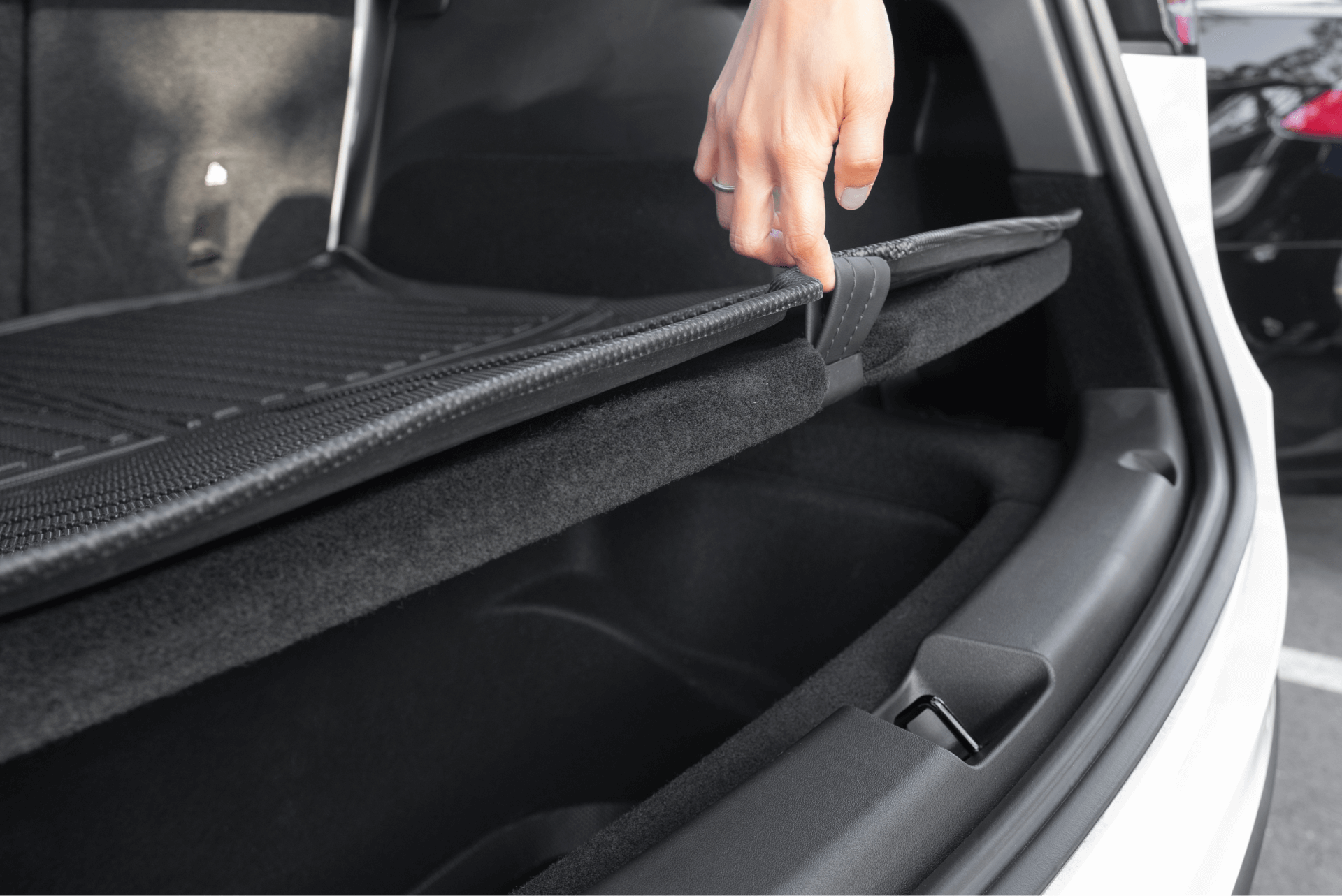 A hand lifts a strap to open a hidden compartment under the trunk floor, revealing extra space ideal for organizing JOWUA Model Y Trunk Combo (2018-2024) accessories or pairing with custom trunk liners.