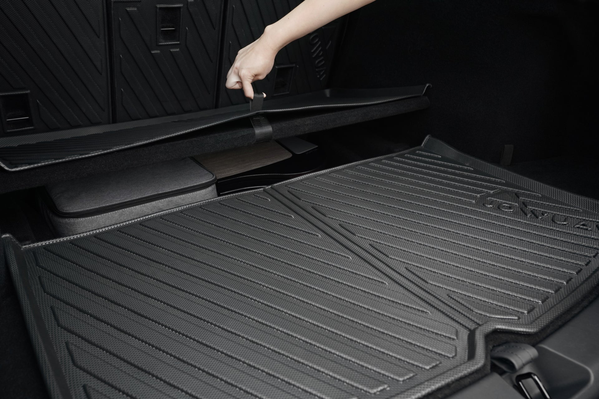 A hand lifts the strap on a Model Y (Juniper 2025+) JOWUA EasyShield Trunk Liner + Rear Seats Back Cover, revealing a storage compartment beneath black, textured waterproof mats.