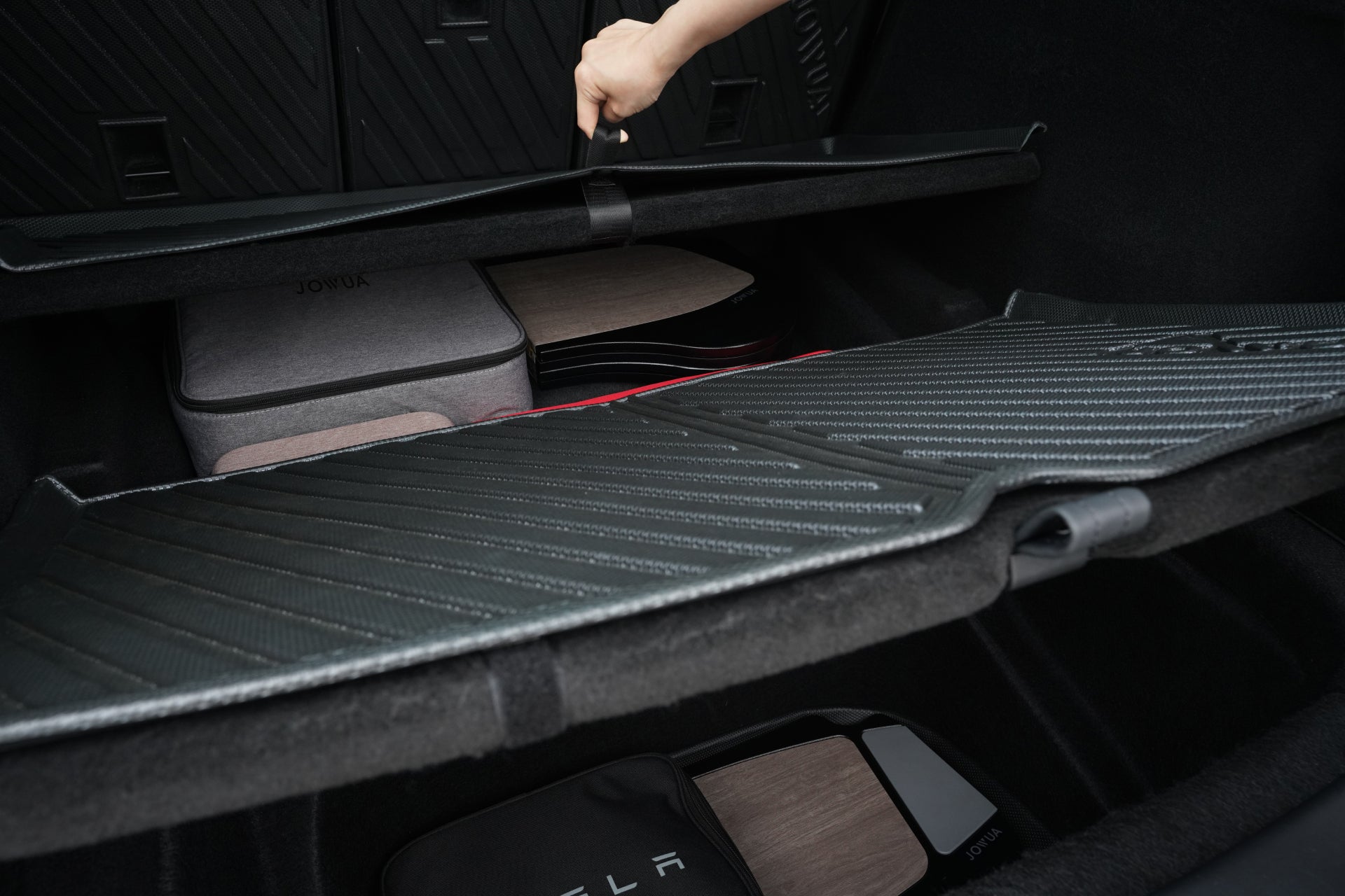 A hand lifts the floor cover in a Tesla Model Y, revealing JOWUA's EasyShield Waterproof Trunk Liner + Rear Seats Back Cover Bundle (Juniper 2025+), with organized storage and Tesla-branded accessories arranged on the premium TPE liner.