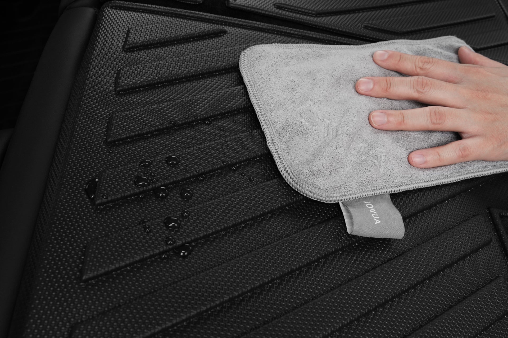 A hand wipes water droplets from a textured black surface, highlighting the JOWUA Model Y (Juniper 2025+) EasyShield Waterproof Trunk Liner + Rear Seats Back Cover Bundle for ultimate Tesla protection.