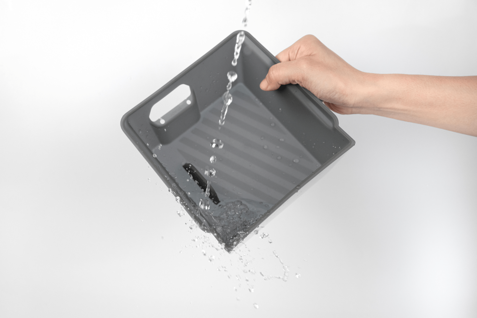 A hand tilts the JOWUA Armrest Organizer Set for Tesla Model Y Juniper 2025+/Model 3 Highland 2024+, made from premium hybrid material, causing water to pour off its grey plastic surface against a plain white background.