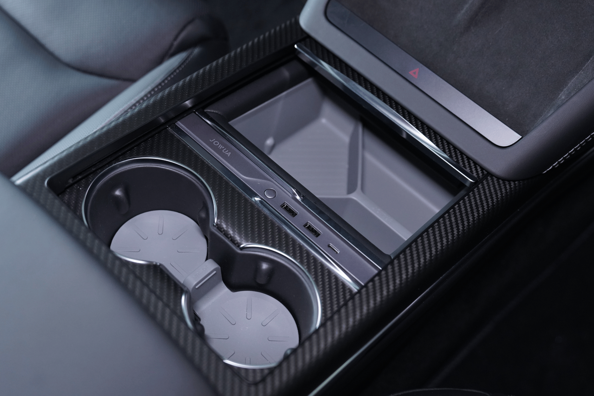The image features a close-up of a car's center console, emphasizing JOWUA's Hub Bar Mini for the Tesla Model S/X. This accessory includes two cup holders and a stylish storage compartment with USB ports. Its modern design, enhanced by a carbon fiber finish and an integrated USB-C hub, exudes minimalistic elegance.