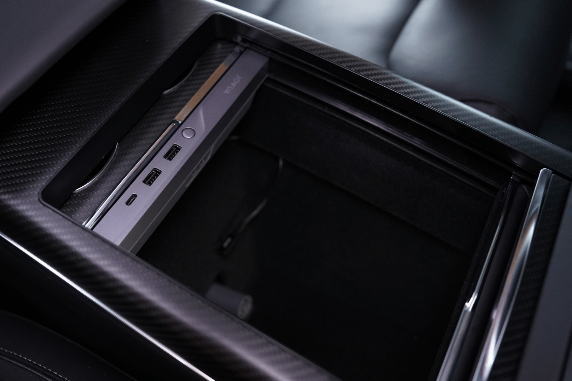 Close-up of the JOWUA Hub Bar Mini for Tesla Model S/X center console compartment, showcasing USB-C hubs and a storage area with a sleek black finish. The product exemplifies spaciousness and modernity, featuring waterproof technology and a textured surface.