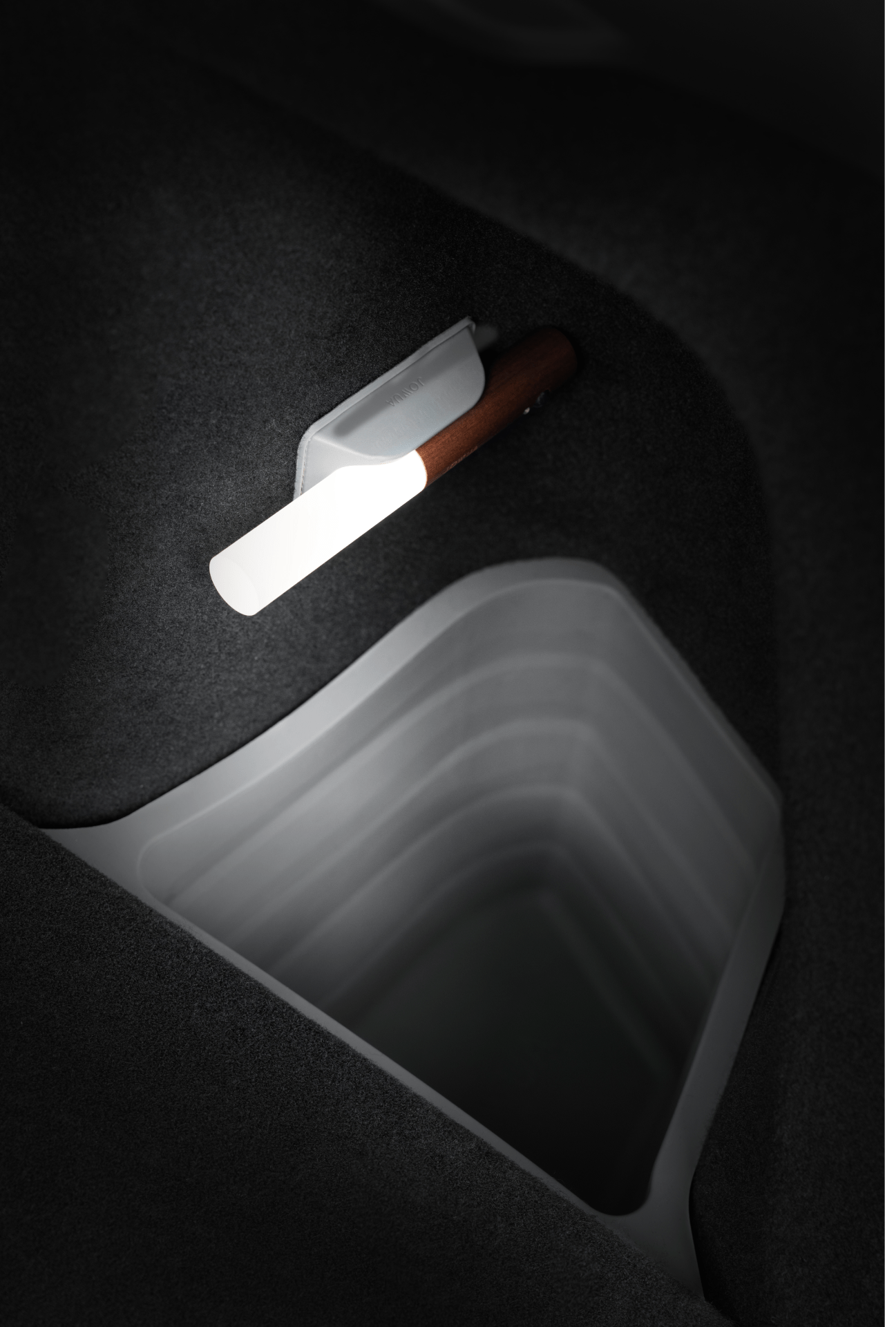 A close-up of a modern wall light with a wooden handle illuminating the JOWUA Trunk Storage Bins Bundle for Model Y (2018-2024), set into a dark, felt-textured surface.