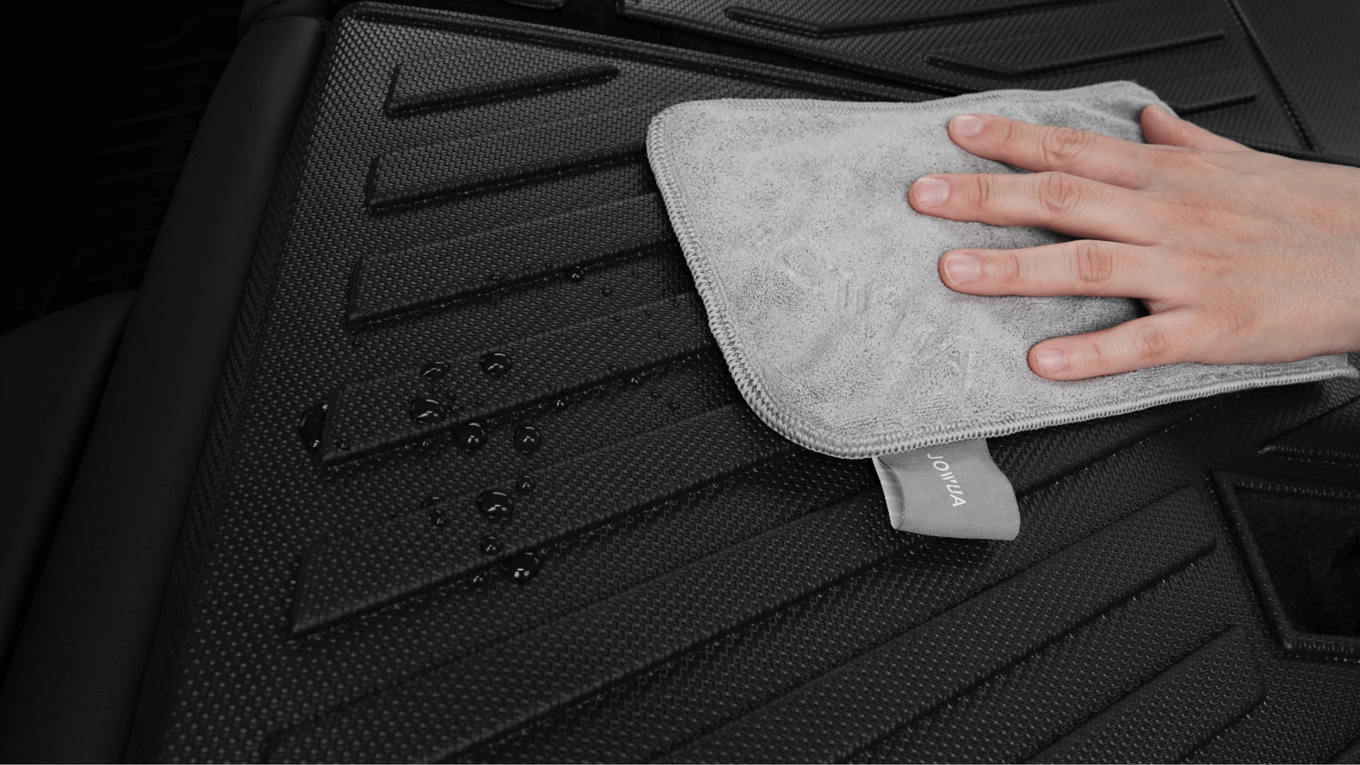 A hand uses a gray microfiber cloth to wipe water droplets off a textured black JOWUA Model Y Trunk Combo (2018-2024) floor mat, an essential accessory for Tesla owners.