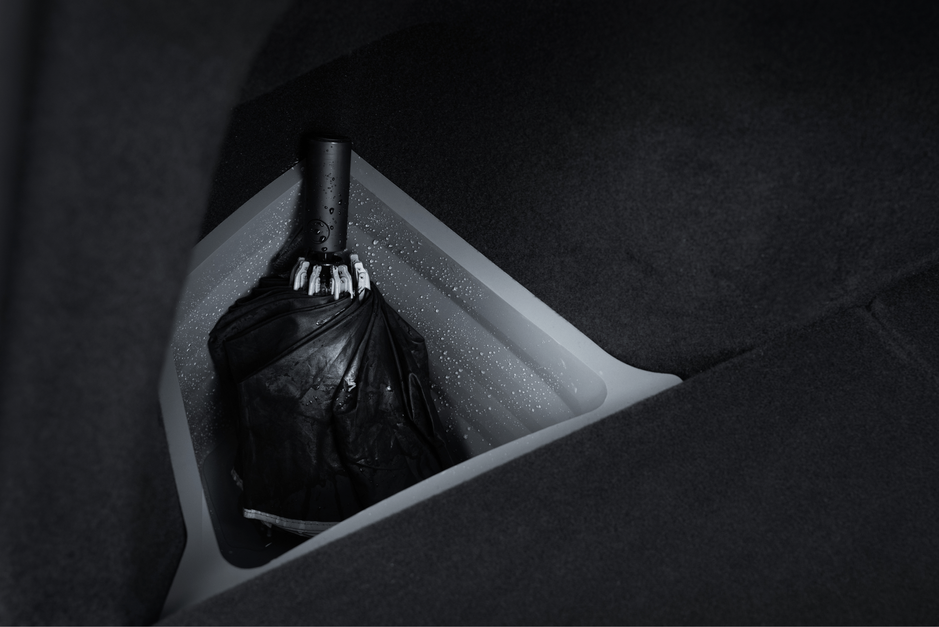 A black, wet umbrella sits in a JOWUA Trunk Storage Bin Bundle for Model Y (2018-2024) with a light grey lining, contrasting with the bin’s dark, soft-textured surfaces. Water droplets appear on the compartment walls.