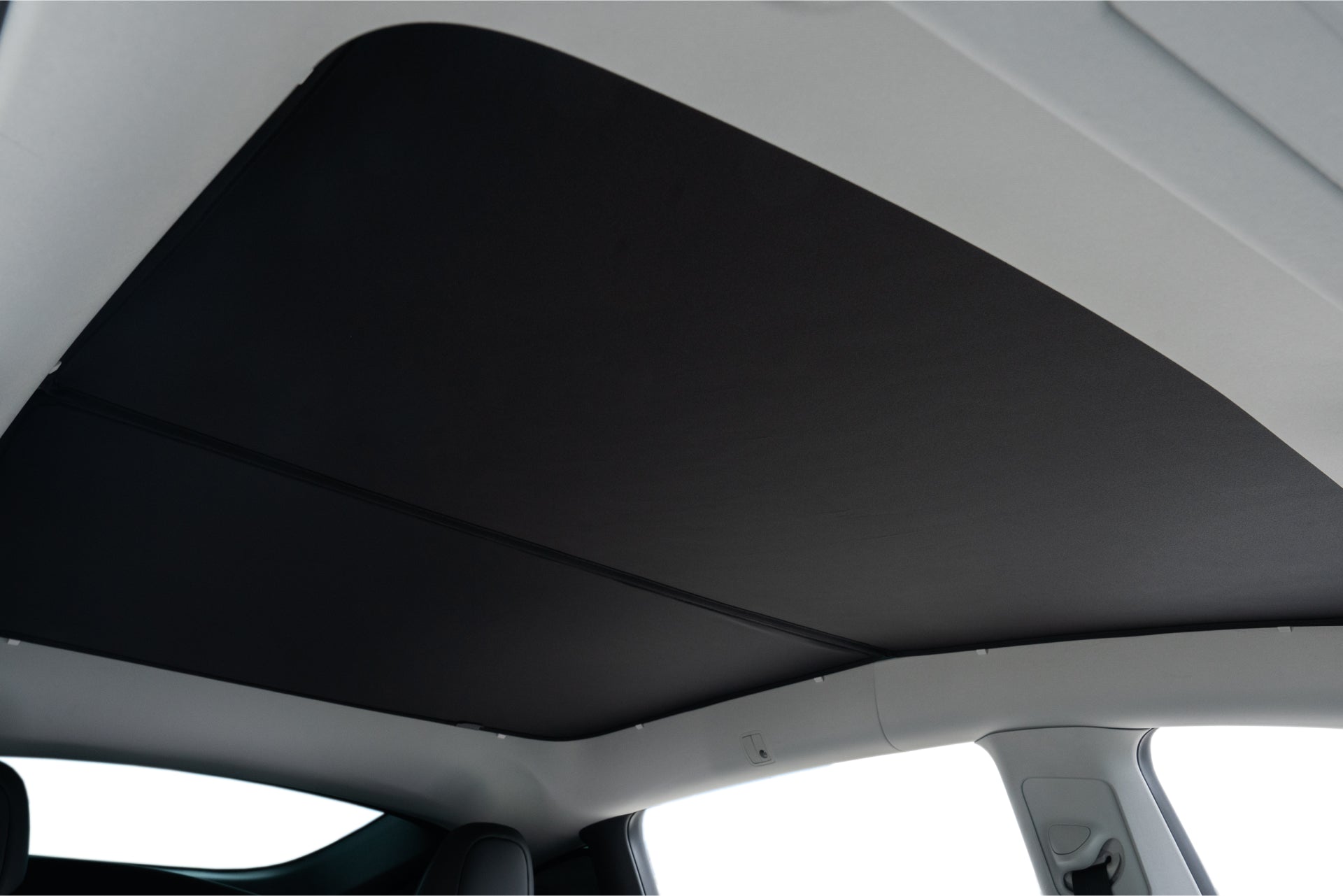 The image showcases the interior roof of a Tesla, highlighting a large panoramic sunroof with tinted glass framed by light-colored trim. The JOWUA Model Y Ultimate Sunshade Bundle offers UV protection, spanning most of the ceiling and providing a spacious and airy feel.