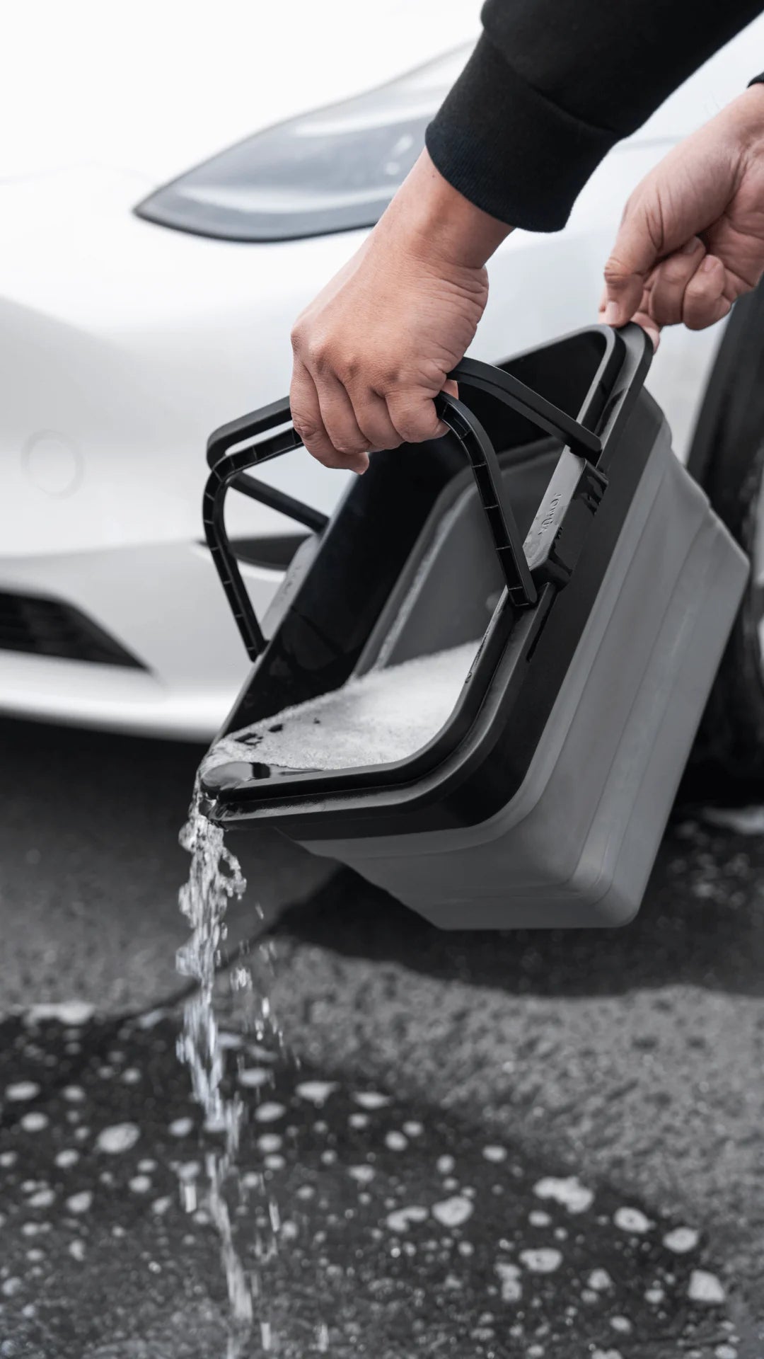 A person is pouring soapy water from a gray bucket onto the pavement, and using the JOWUA Microfiber Cleaning Cloth from the Under Seat Collapsible Organizer & Microfiber Cleaning Cloth Combo for a shiny finish. In the background, part of a white car gleams under the sunlight. The soapy water splashes as it hits the ground, creating bubbles that catch reflections.