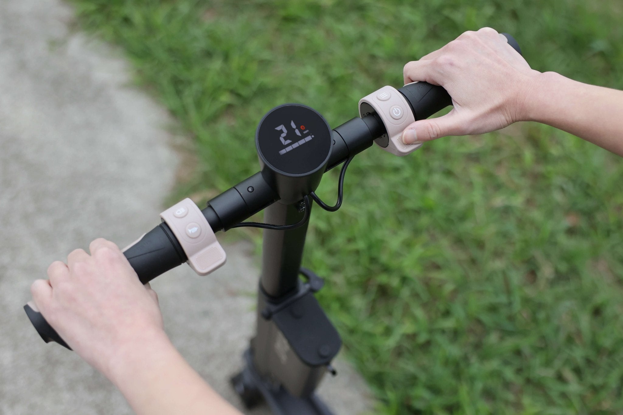 Hands are gripping the handlebar of JOWUA's Folding Electric Scooter (Long Range) for Tesla (S3XY) on a path. The digital display indicates the speed and battery level, with green grass in the background. Ideal for city commuting, its detachable battery offers convenience and ease of use.