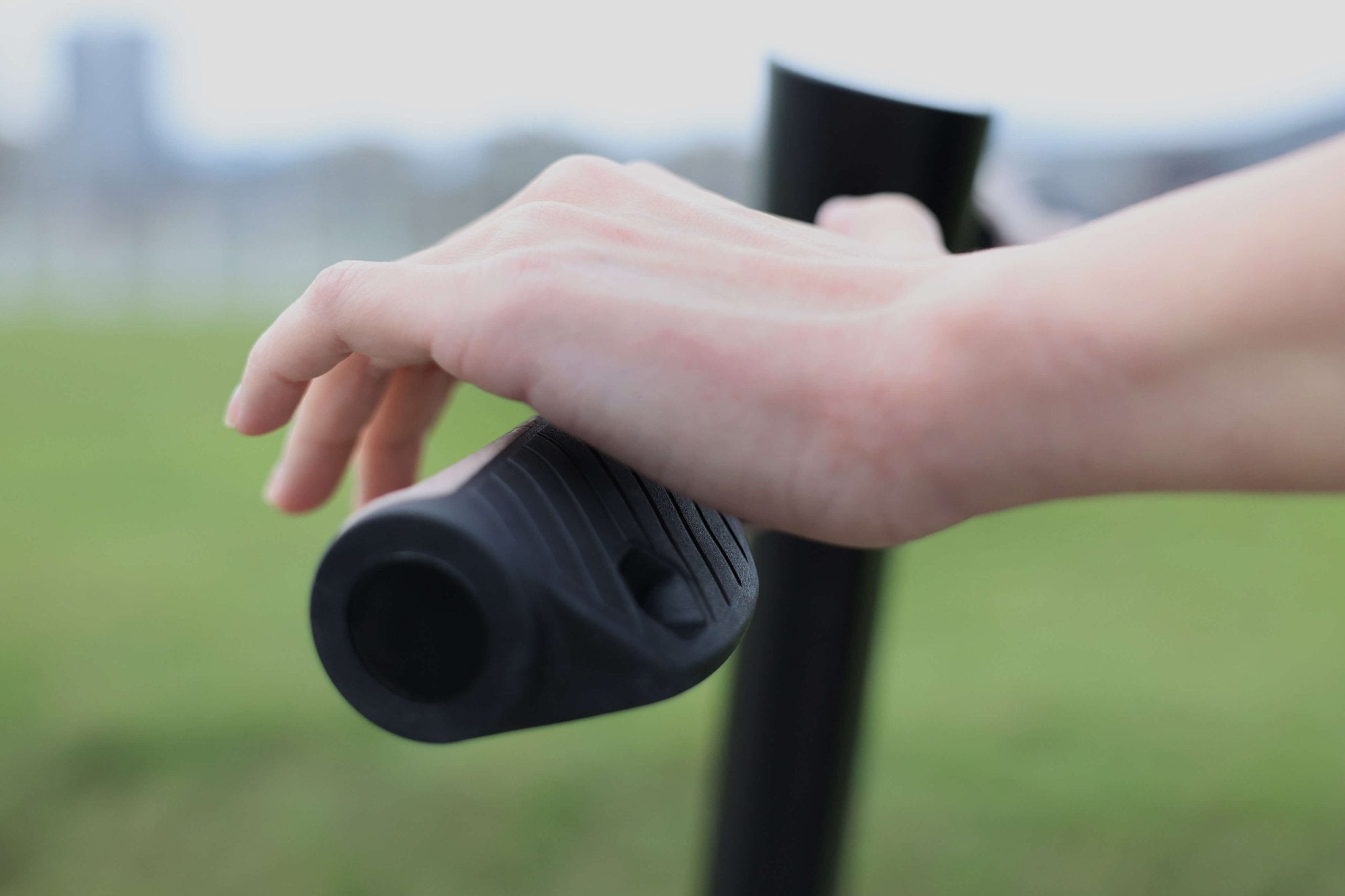 A close-up of a hand gently gripping the handle of the JOWUA Folding Electric Scooter (Long Range) for Tesla (S3XY), with a blurred background of green grass and blue sky, reminiscent of securing an anti-theft padlock to ensure peace of mind.