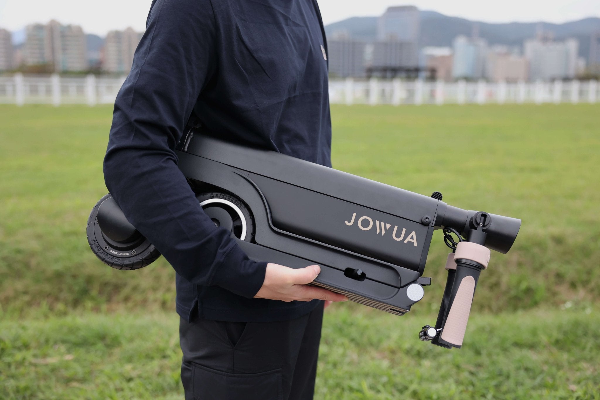 A person in a dark sweatshirt holds a folded JOWUA folding electric scooter, designed for long range and Tesla (S3XY), showcasing its sleek design and detachable battery. They stand on green grass with city buildings in the background.