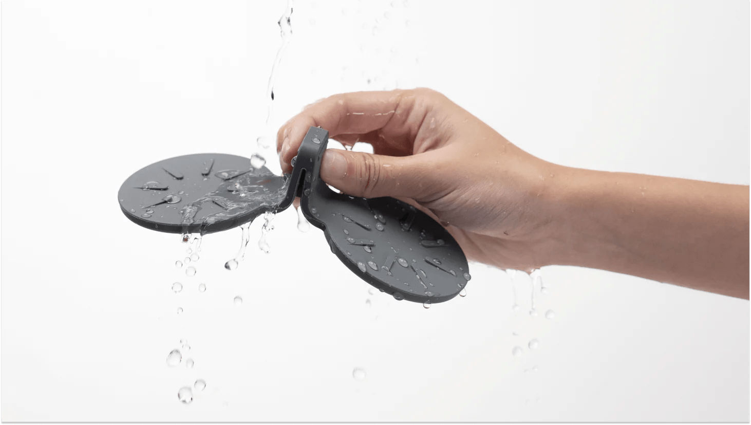 A person's hand holds a dark gray, water-resistant Rear Seat Coaster for Tesla Model S by JOWUA, crafted from premium silicone with two circular pads connected by a central piece. Water droplets cascade over the coaster and hand against a plain white background, highlighting its non-slip design.