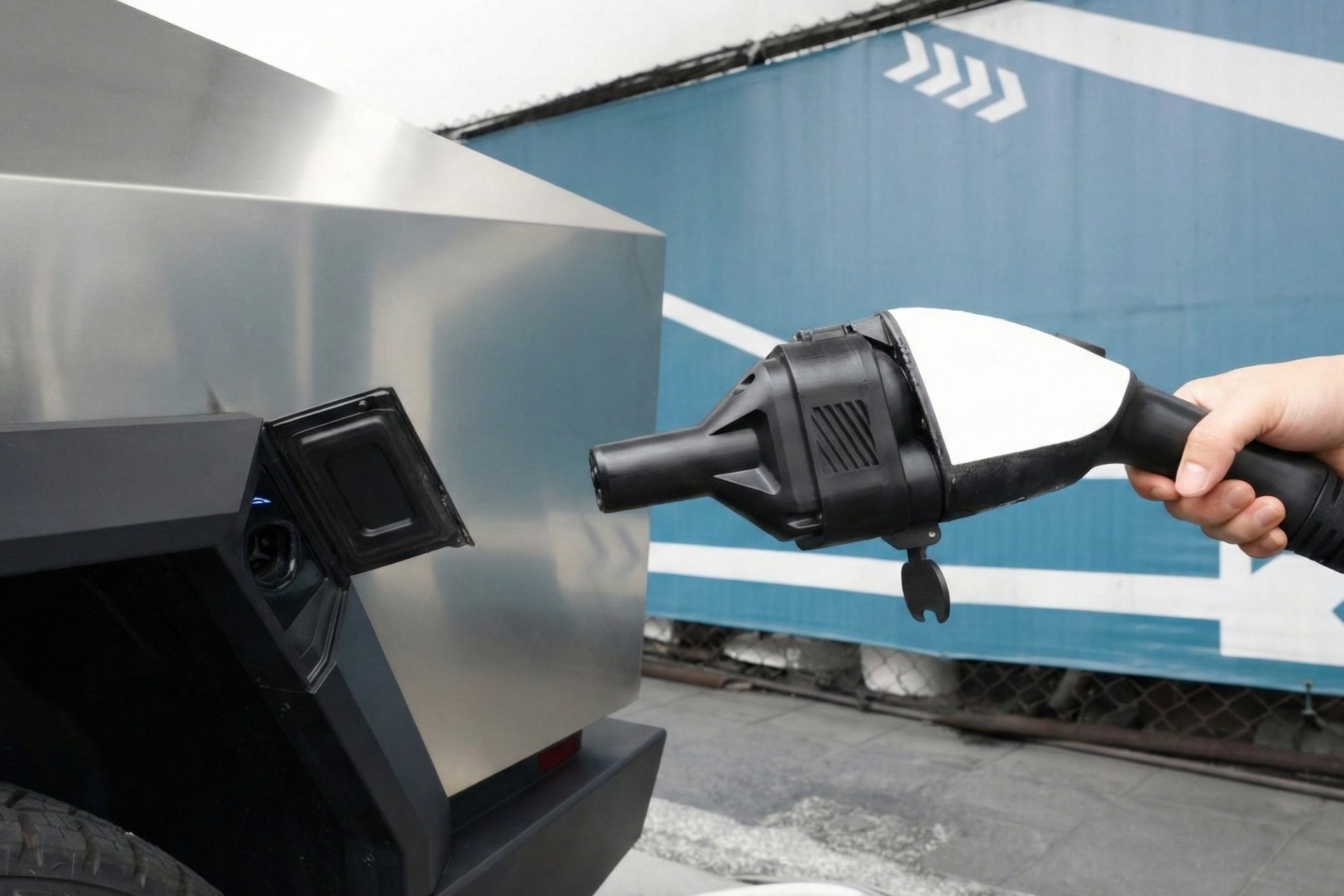 A person holds a JOWUA CCS1 to NACS DC & AC Charging Adapter near the charging port of a silver, angular electric truck, with a blue wall featuring white arrow graphics in the background.