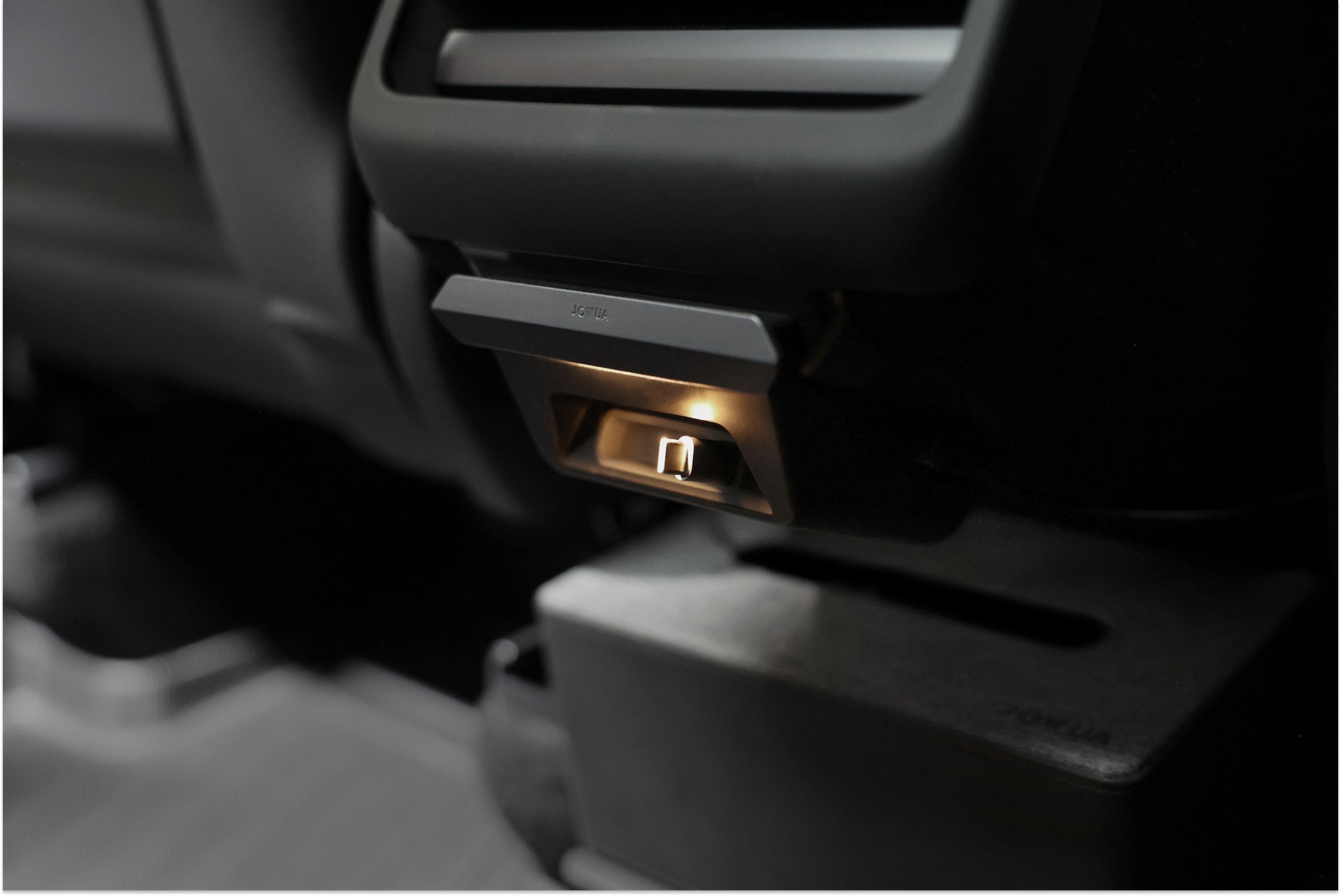 Close-up of a JOWUA 130W LED Rear Vent Hub (Model 3 Highland 2026+/Model Y Juniper 2026+) illuminating an interior latch, with dark plastic parts and textured flooring visible under a seat or dashboard.
