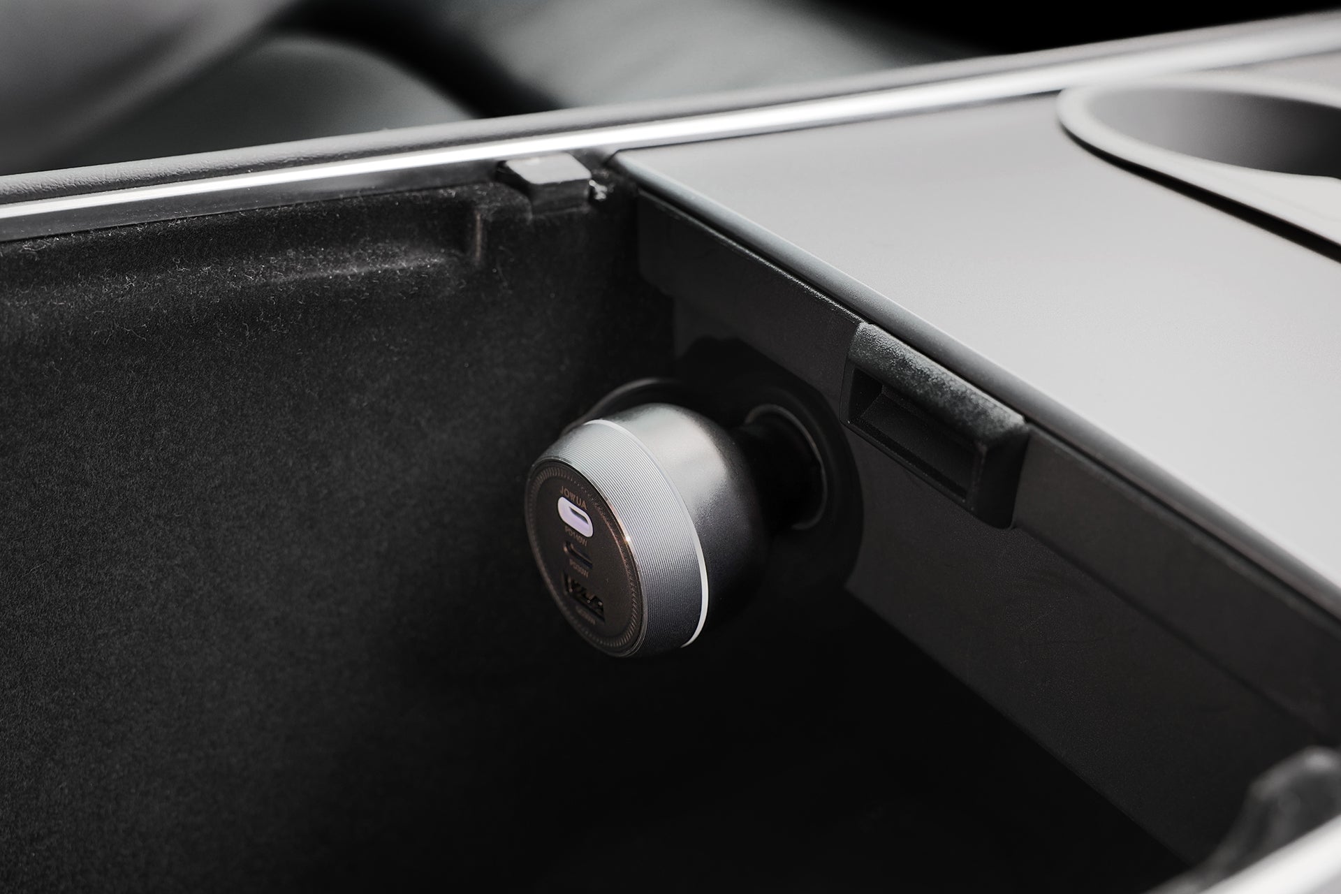 A close-up of a JOWUA Portable Air Compressor + Charging Solution Kit in a car’s center console, showing its round USB high-speed charging port and an adjacent empty cup holder in a clean, modern interior.