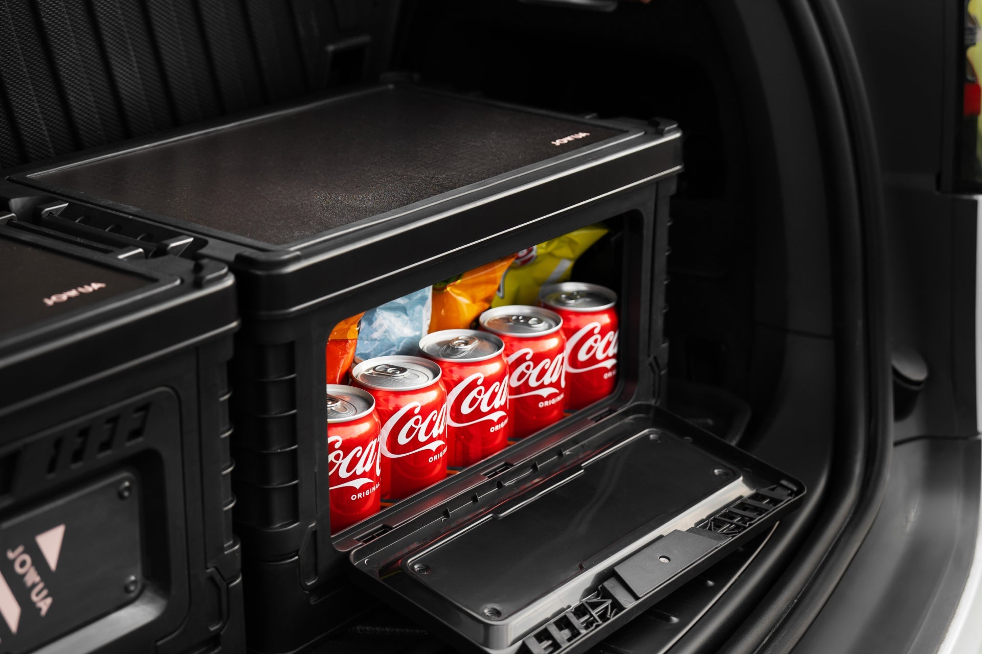 A JOWUA 30L Stackable Storage Box and Trunk Organizer for Teslas sits open in a car trunk, displaying neatly arranged cans of Coca-Cola and bags of snacks inside.
