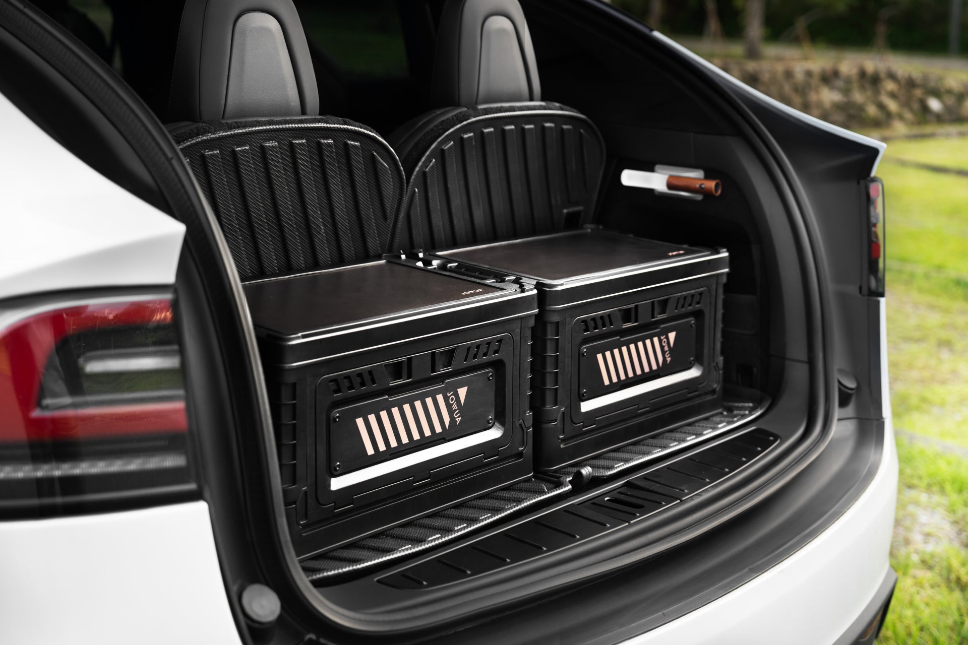 Two large black JOWUA 30L Stackable Storage Box and Trunk Organizers for Teslas are placed in the rear cargo area of an electric vehicle with the hatch open, showing the back seats and outdoor scenery in the background.