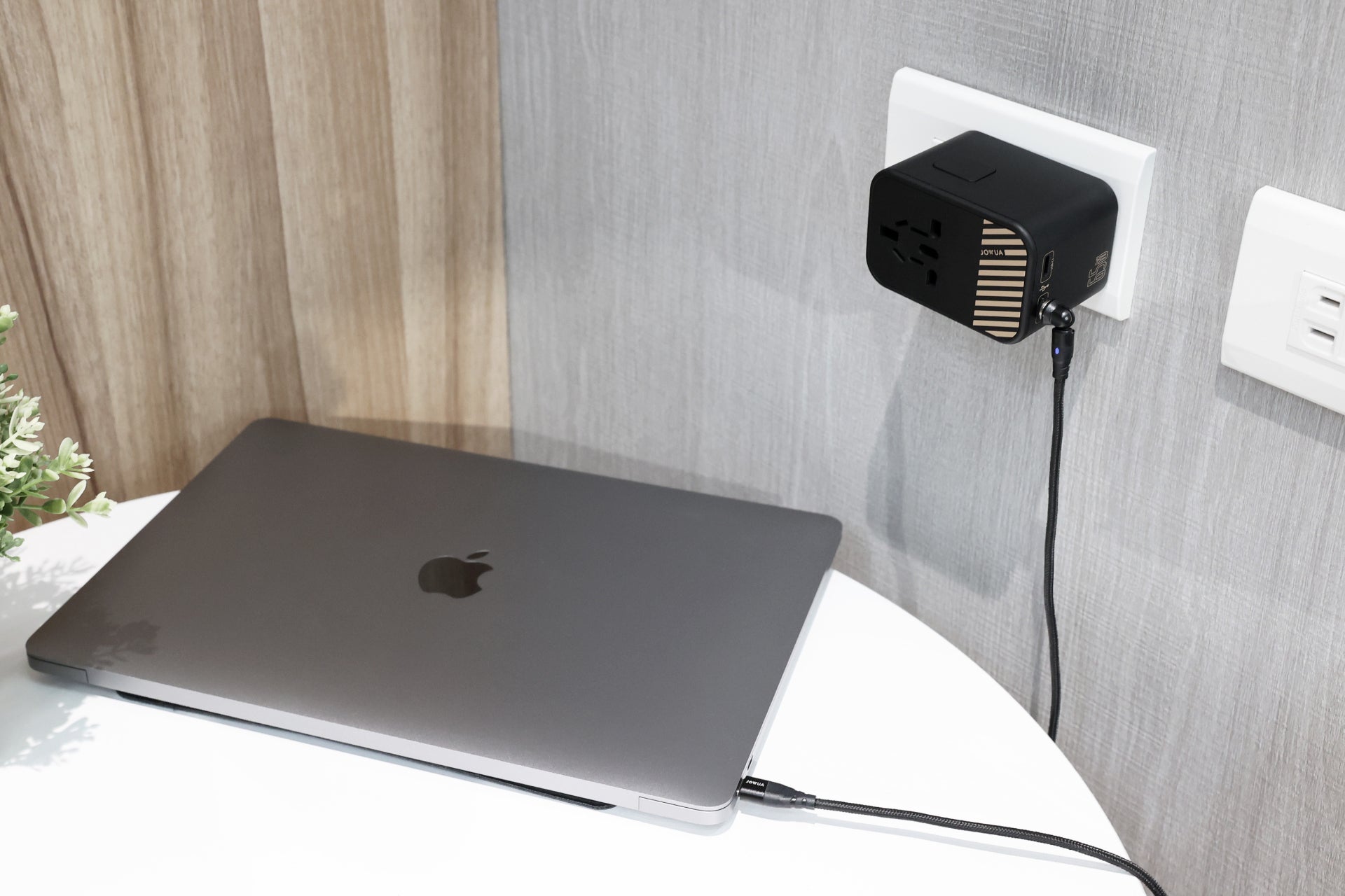 A closed MacBook sits on a white table, connected to the JOWUA 65W GaN Travel Adapter plugged into an outlet on a gray wall. A small plant on the left highlights the adapter’s convenient international charging capabilities.