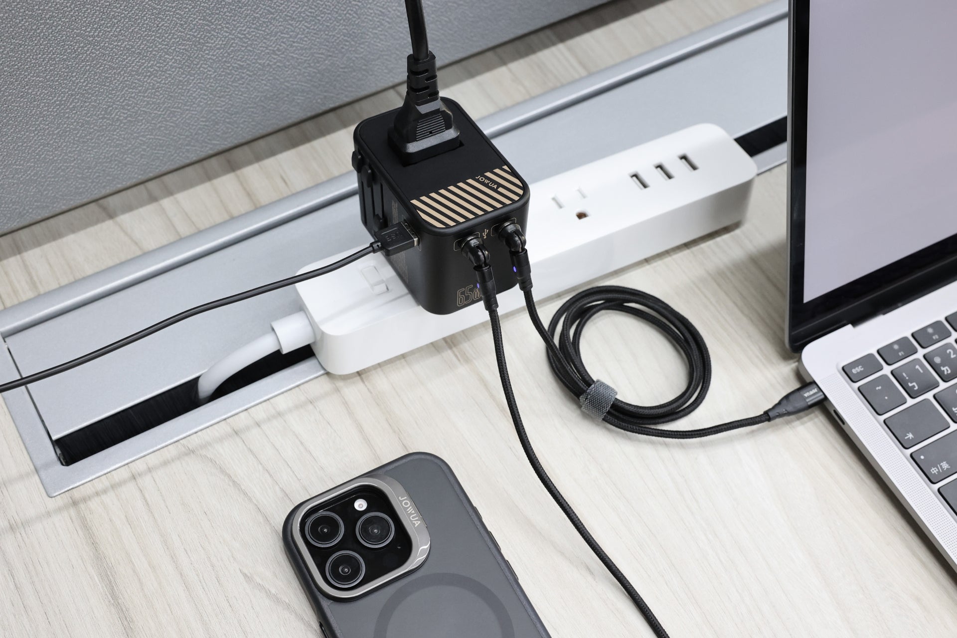 A JOWUA 65W GaN Travel Adapter sits on a light desk, with a smartphone face up beside a laptop and multiple cables connected. Its multiple USB and plug-in ports enable fast charging for all devices.